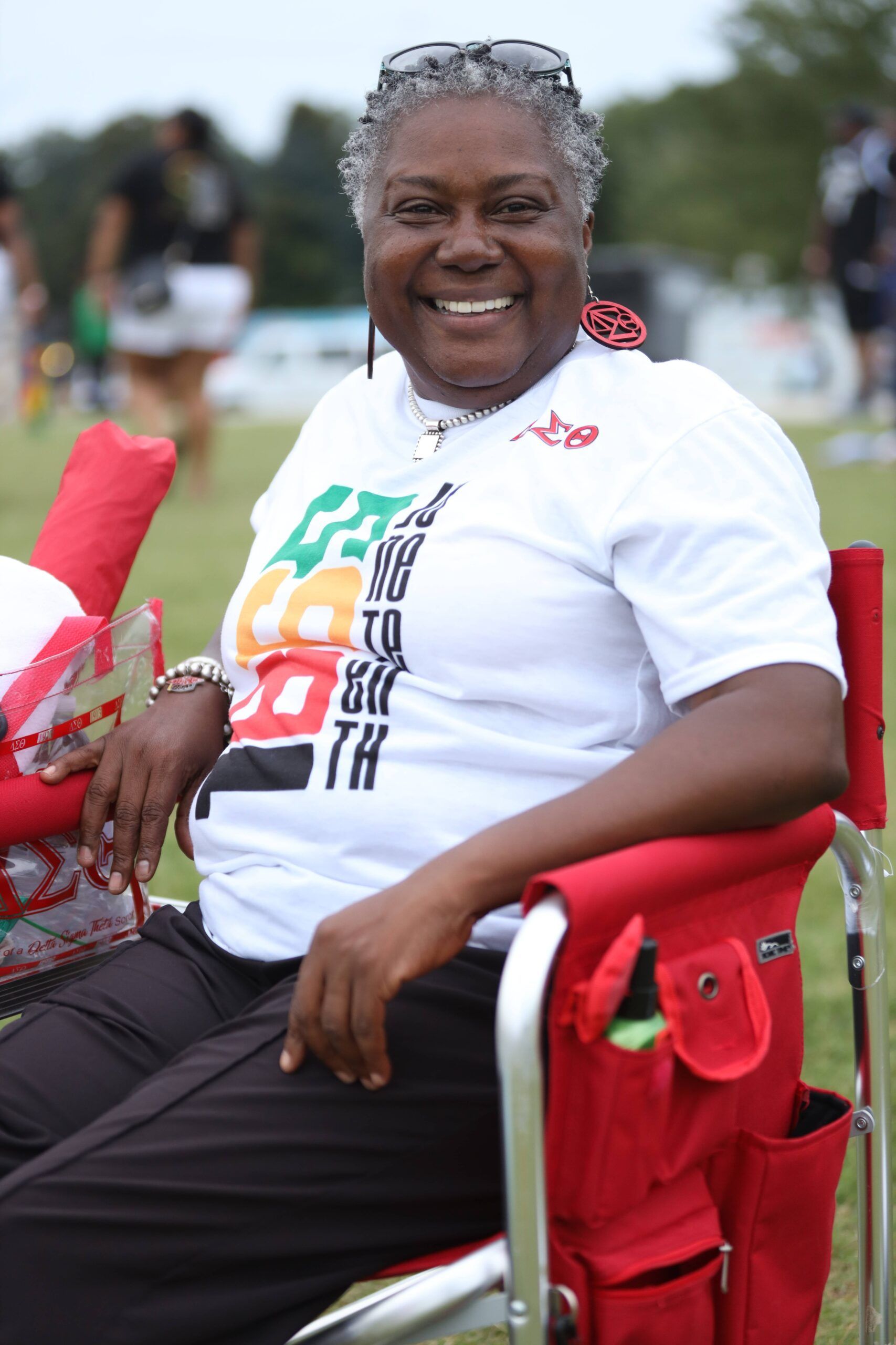 Smiling person seated outdoors in a red folding chair, wearing a white T-shirt and black pants.