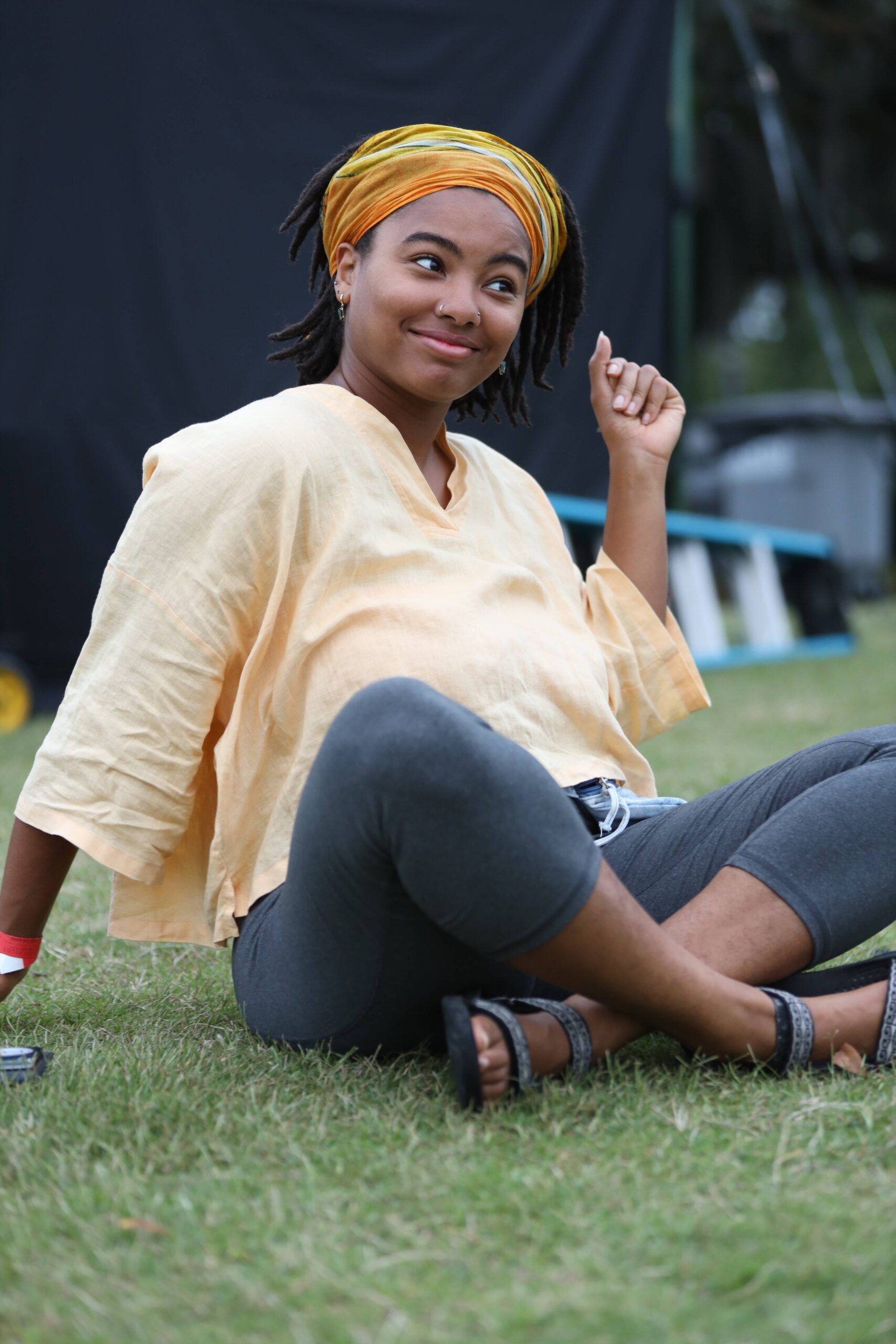 Woman sitting on grass, smiling and holding up a finger, wearing a yellow headwrap and loose shirt.