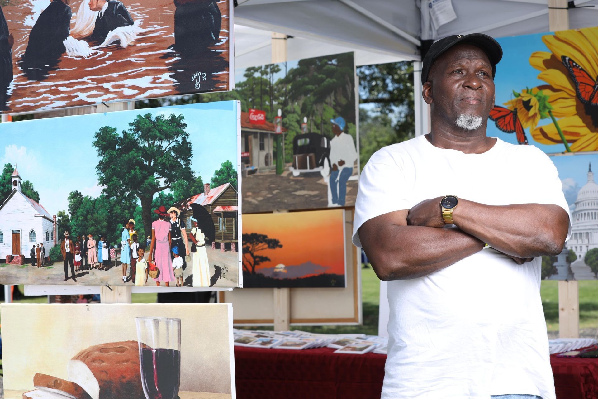 Man in white T-shirt and cap standing with arms crossed at an outdoor event display of photos and paintings