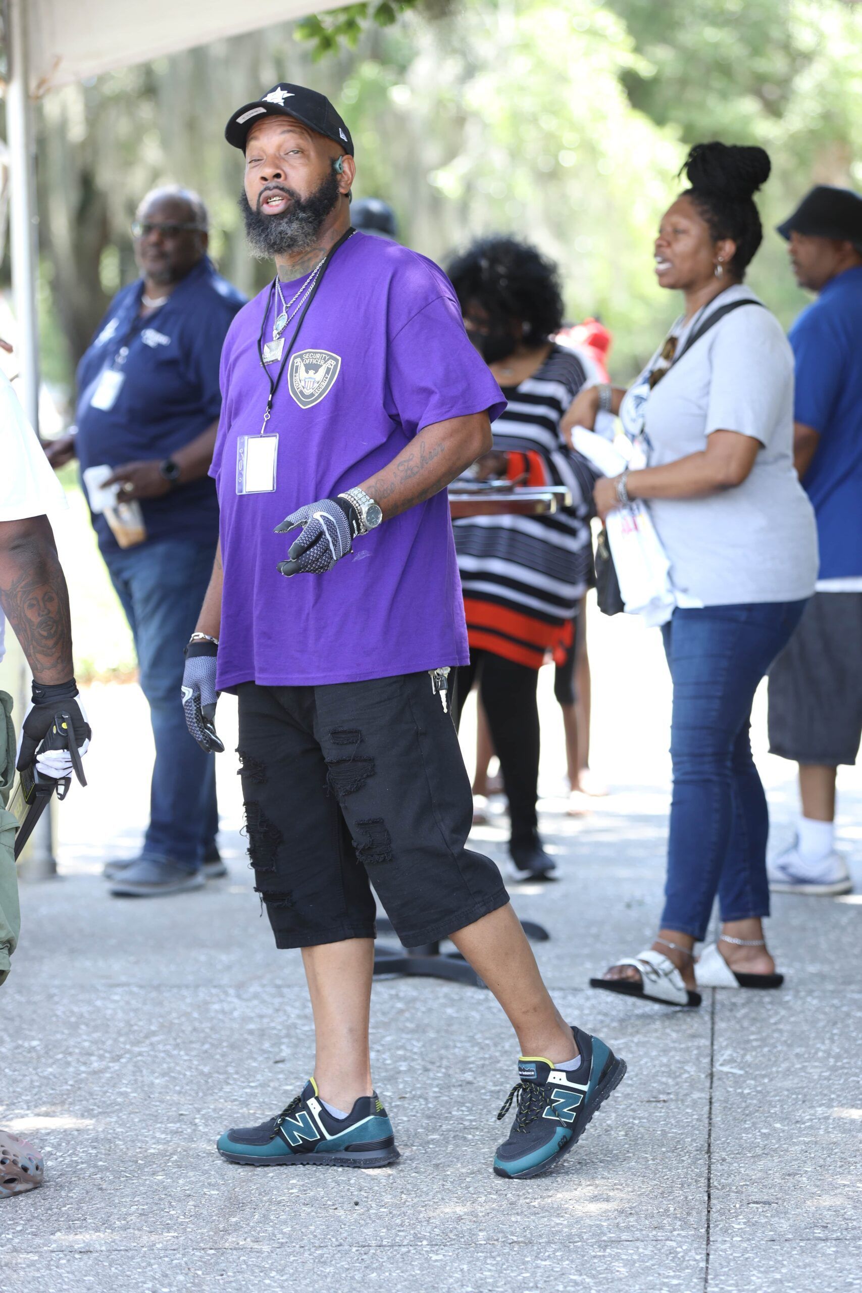 Man in purple shirt standing outdoors among a small crowd, holding a white cup.