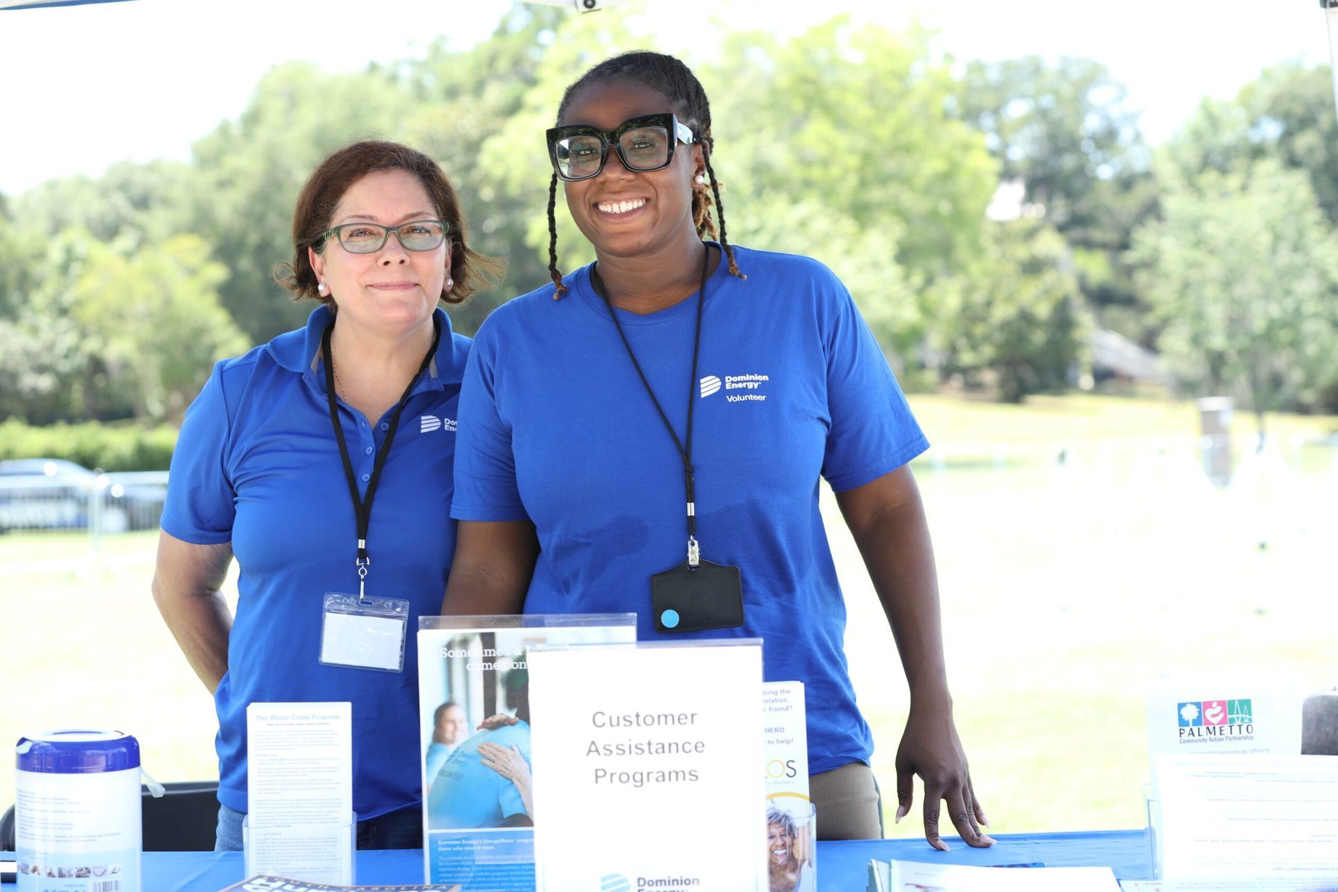 Two volunteers in blue shirts stand behind a table outdoors with informational materials.
