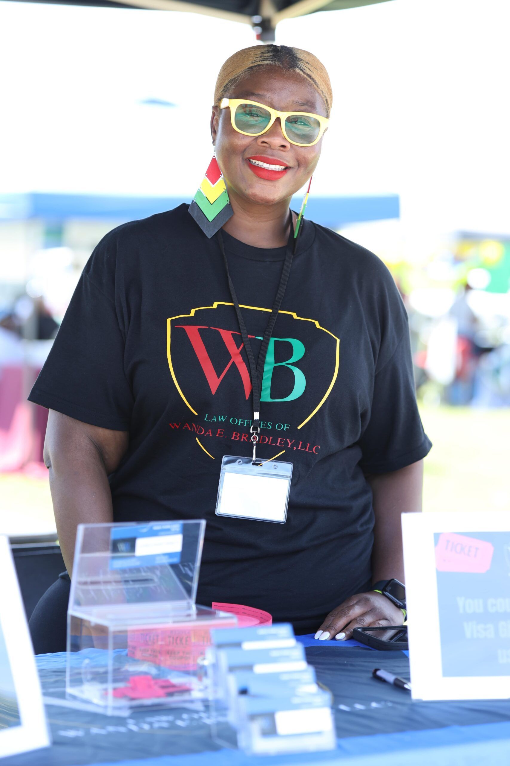 Vendor smiling at a table, wearing a black “WB” shirt, colorful earrings, and neon sunglasses.