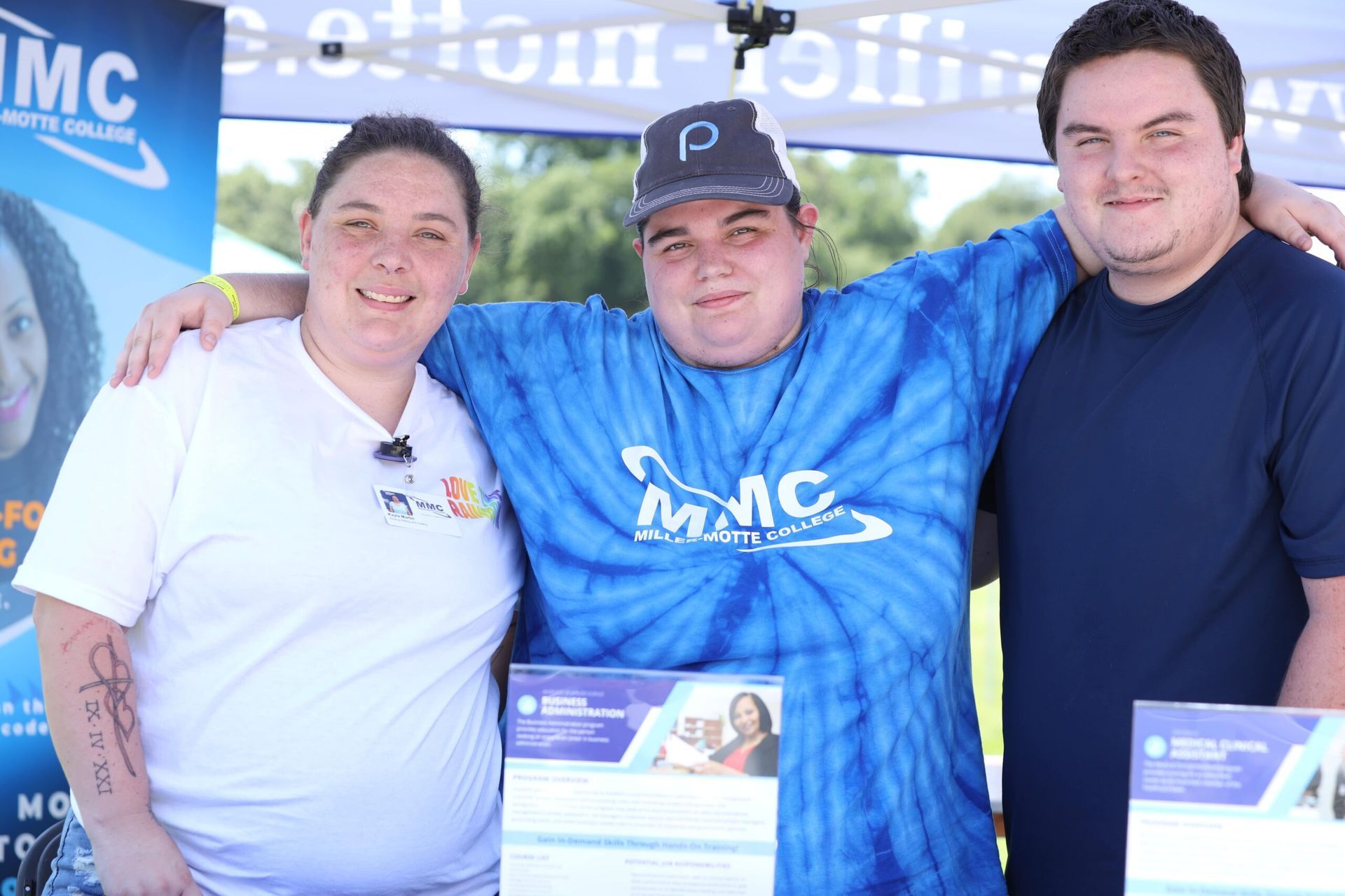 Three people posing at an outdoor MNC event, wearing branded shirts and holding flyers.