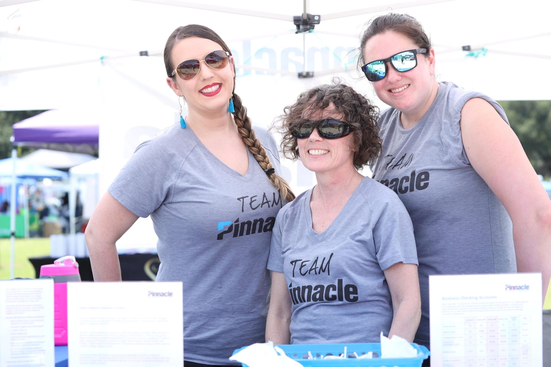 Three people in matching gray shirts smiling behind a table at an outdoor event.