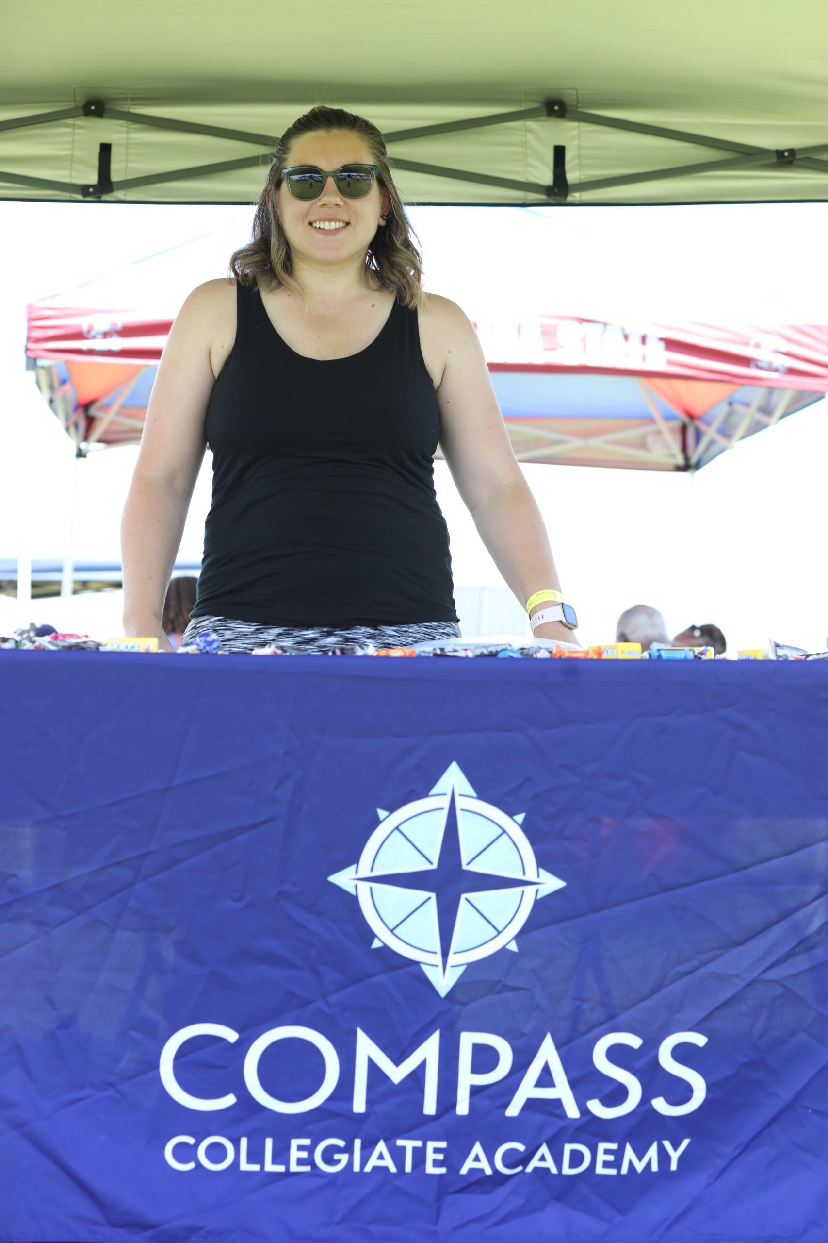 Woman standing behind a blue Compass Collegiate Academy banner under a green canopy.