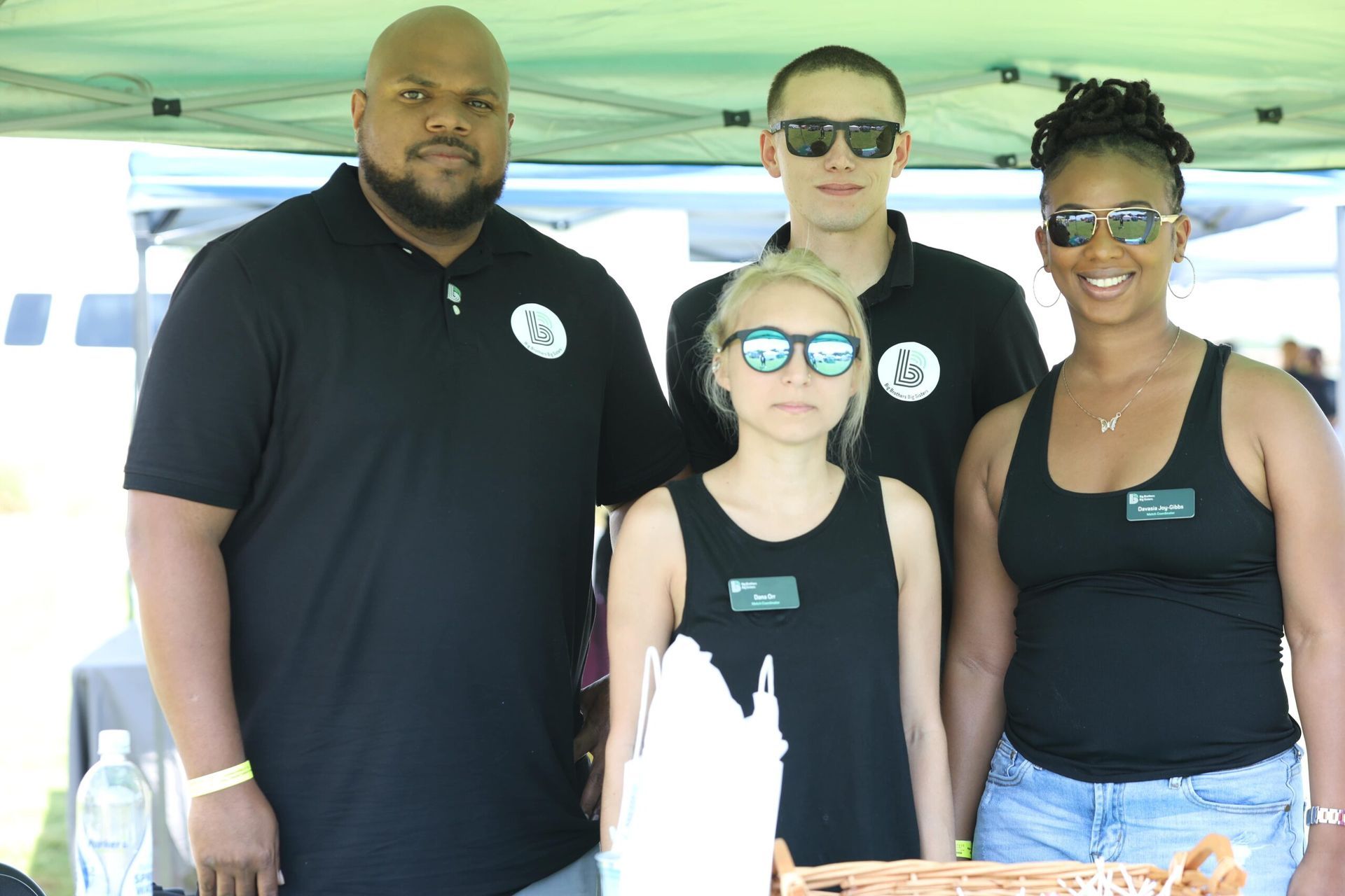 Four people in black shirts standing under a canopy, posing together outdoors with sunglasses on.