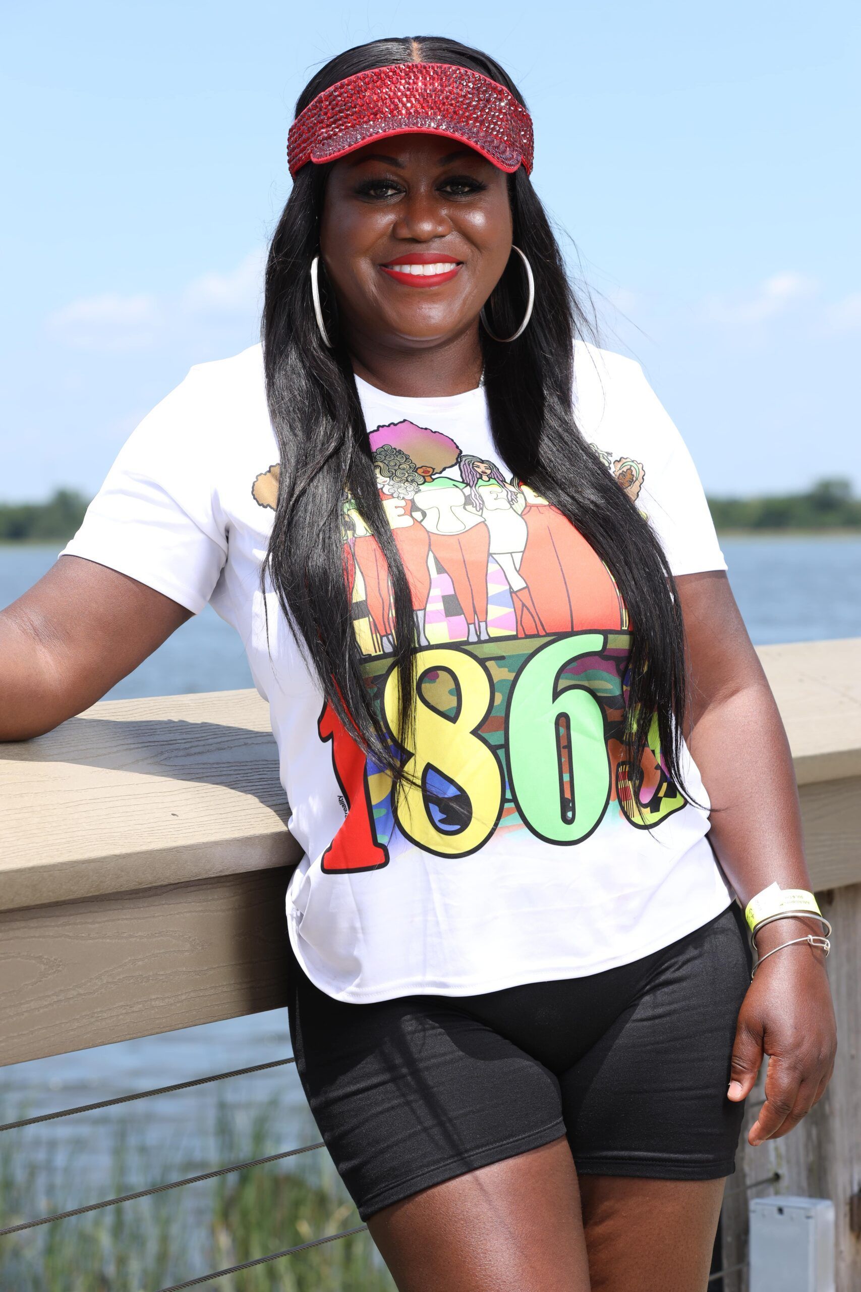 Woman in white graphic T-shirt, black shorts and red cap, smiling by a waterfront railing.