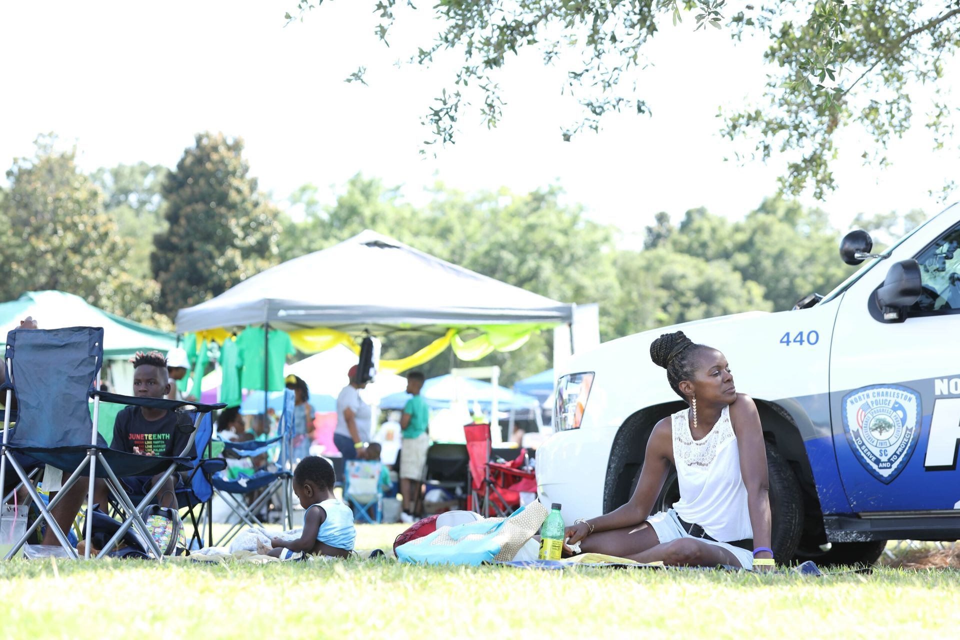 People resting under tents in a park beside a police vehicle on a sunny day