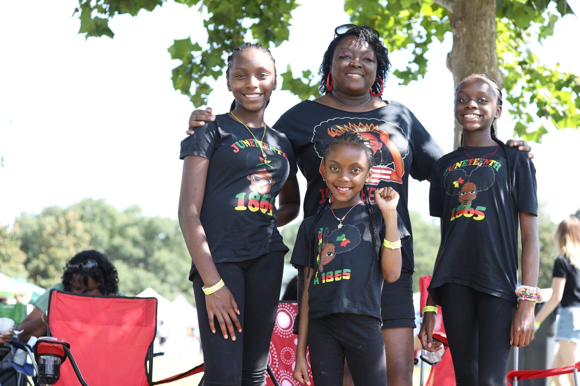 Four people in matching black shirts standing outdoors, smiling with trees and red chairs behind them.