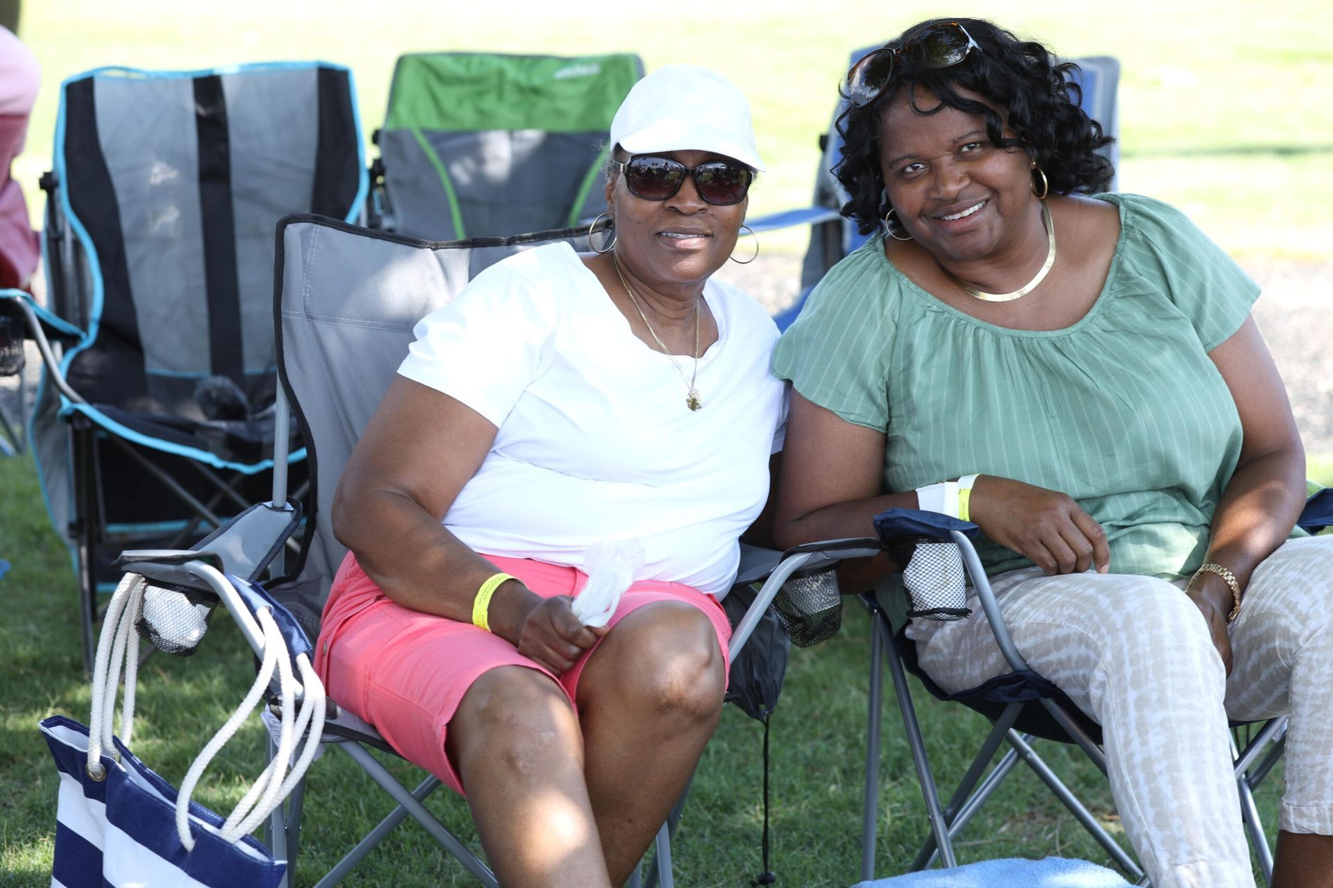 Two women smiling in folding chairs outdoors, one in a white shirt and pink shorts, the other in a green top.