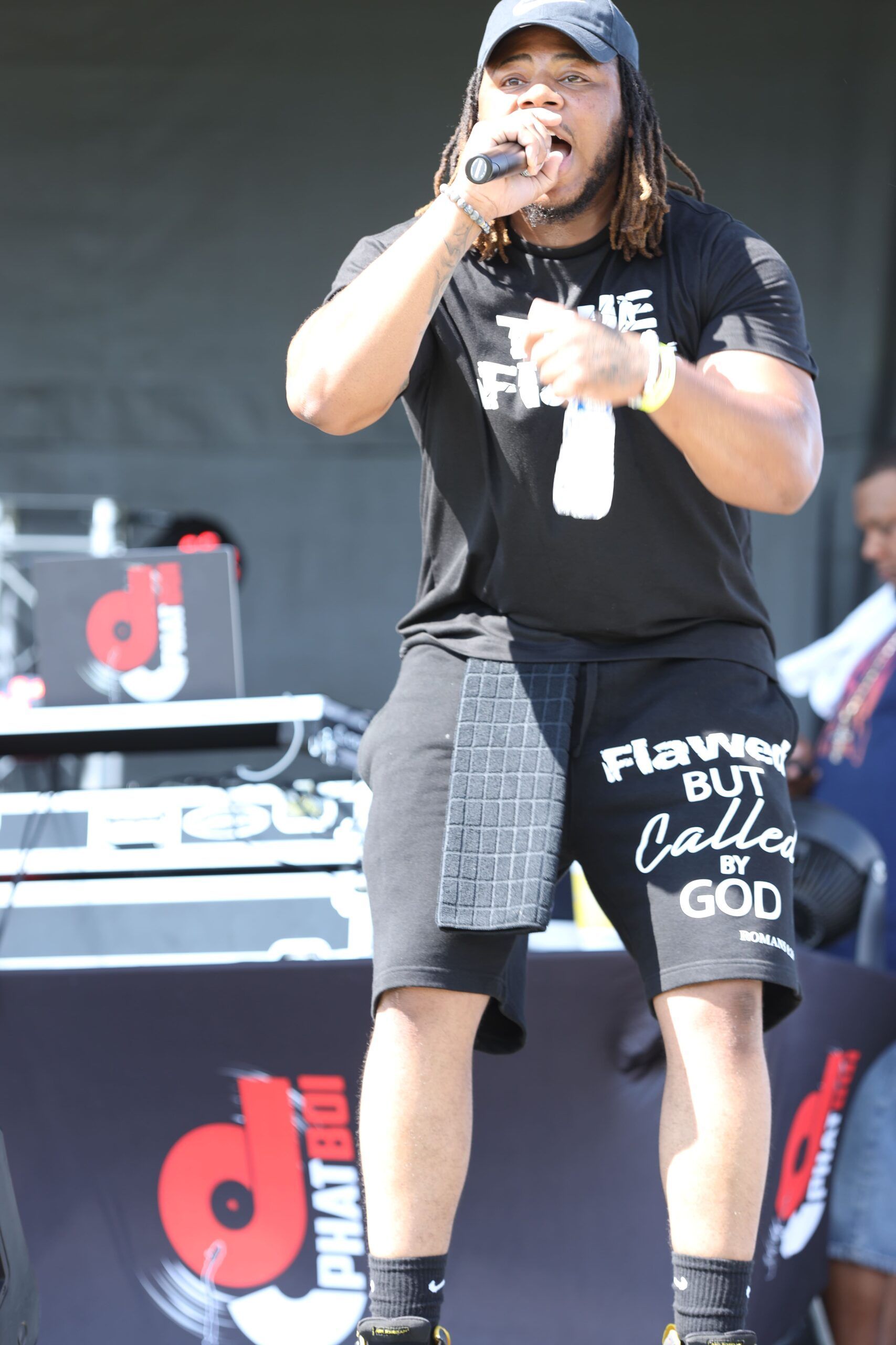 Singer performing onstage in a black outfit and cap, holding a microphone with one hand raised.