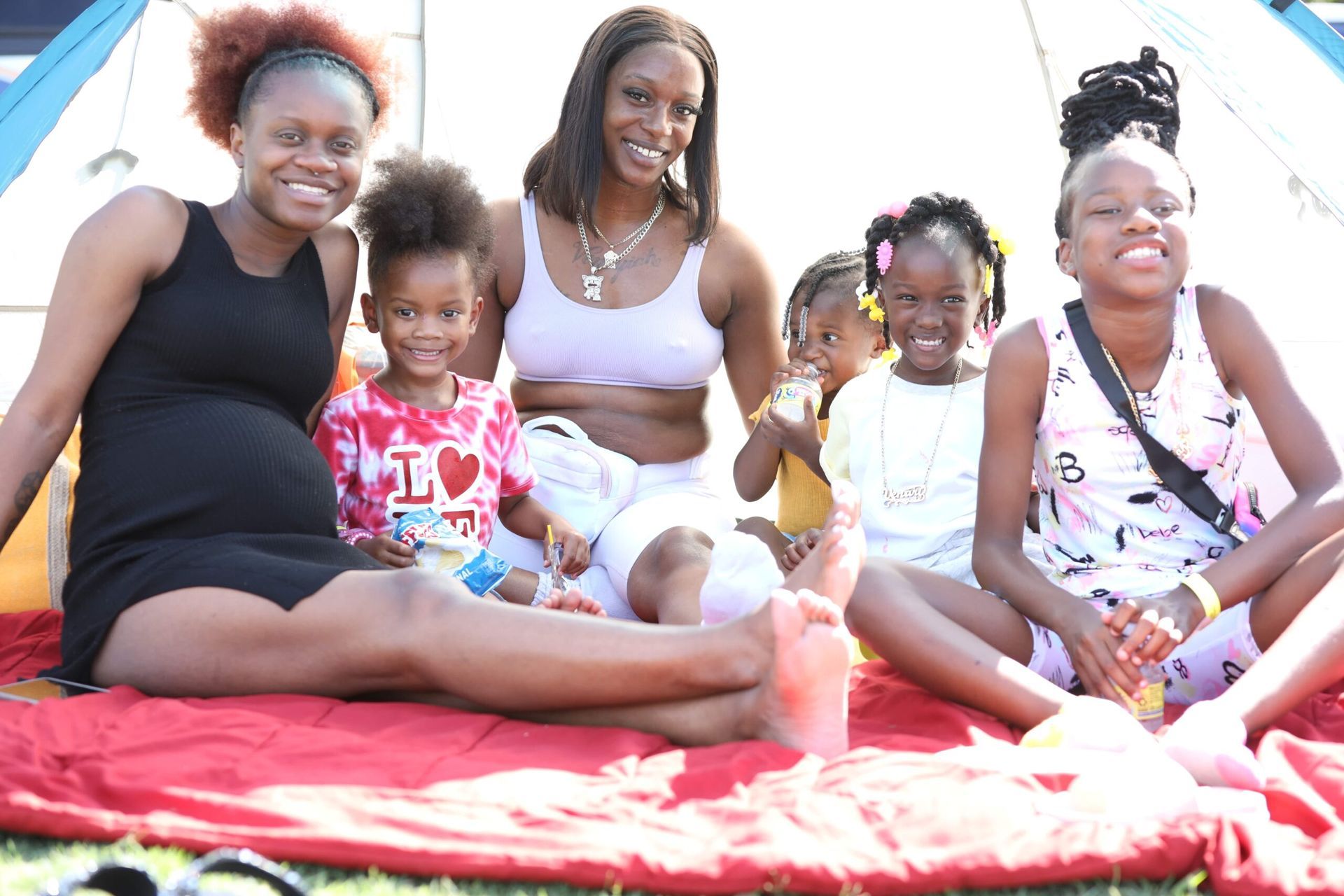 Five people sitting on a red blanket outdoors, smiling at the camera, with a small child holding a doll.