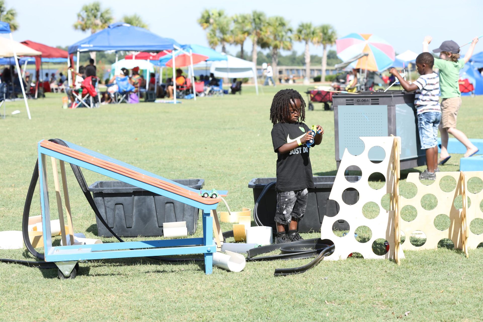 Children playing with outdoor games on a grassy field at a sunny park, with tents and umbrellas in the background