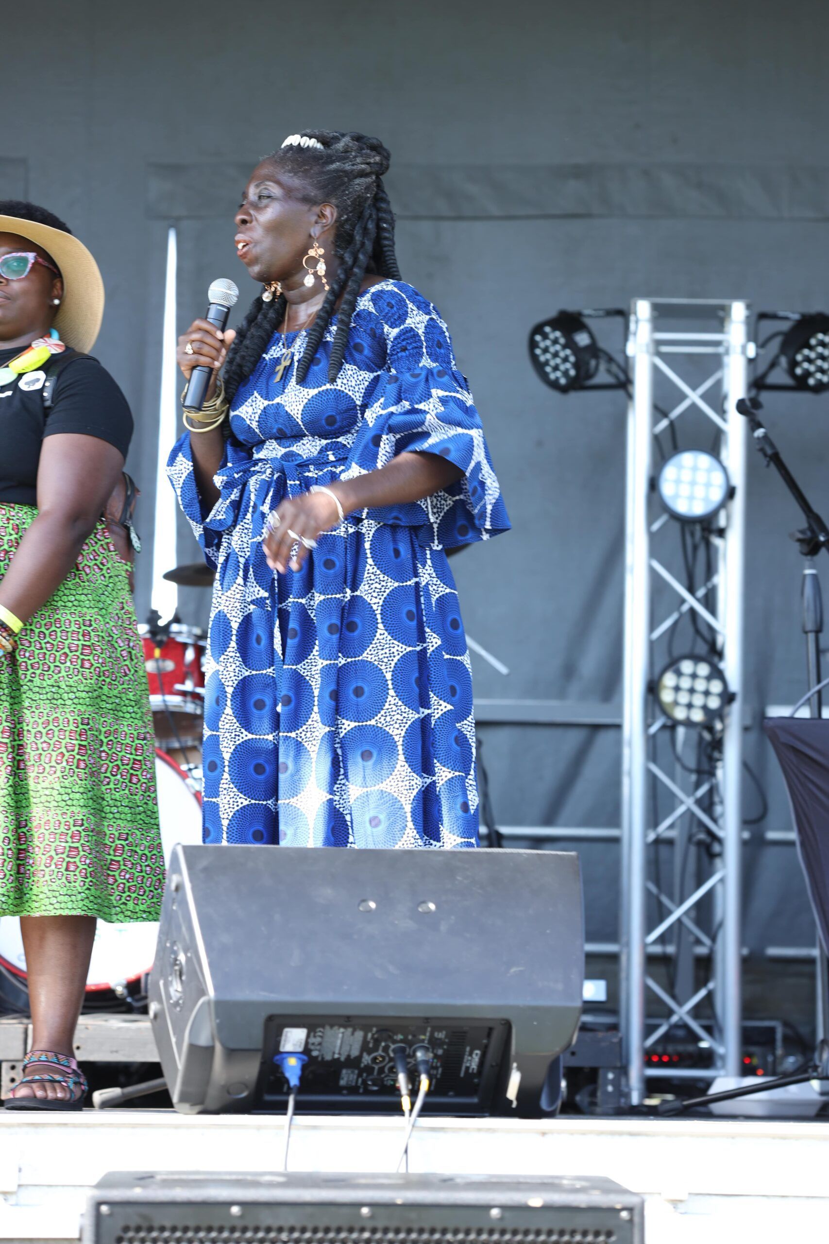 Woman in a blue patterned dress speaking into a microphone on an outdoor stage