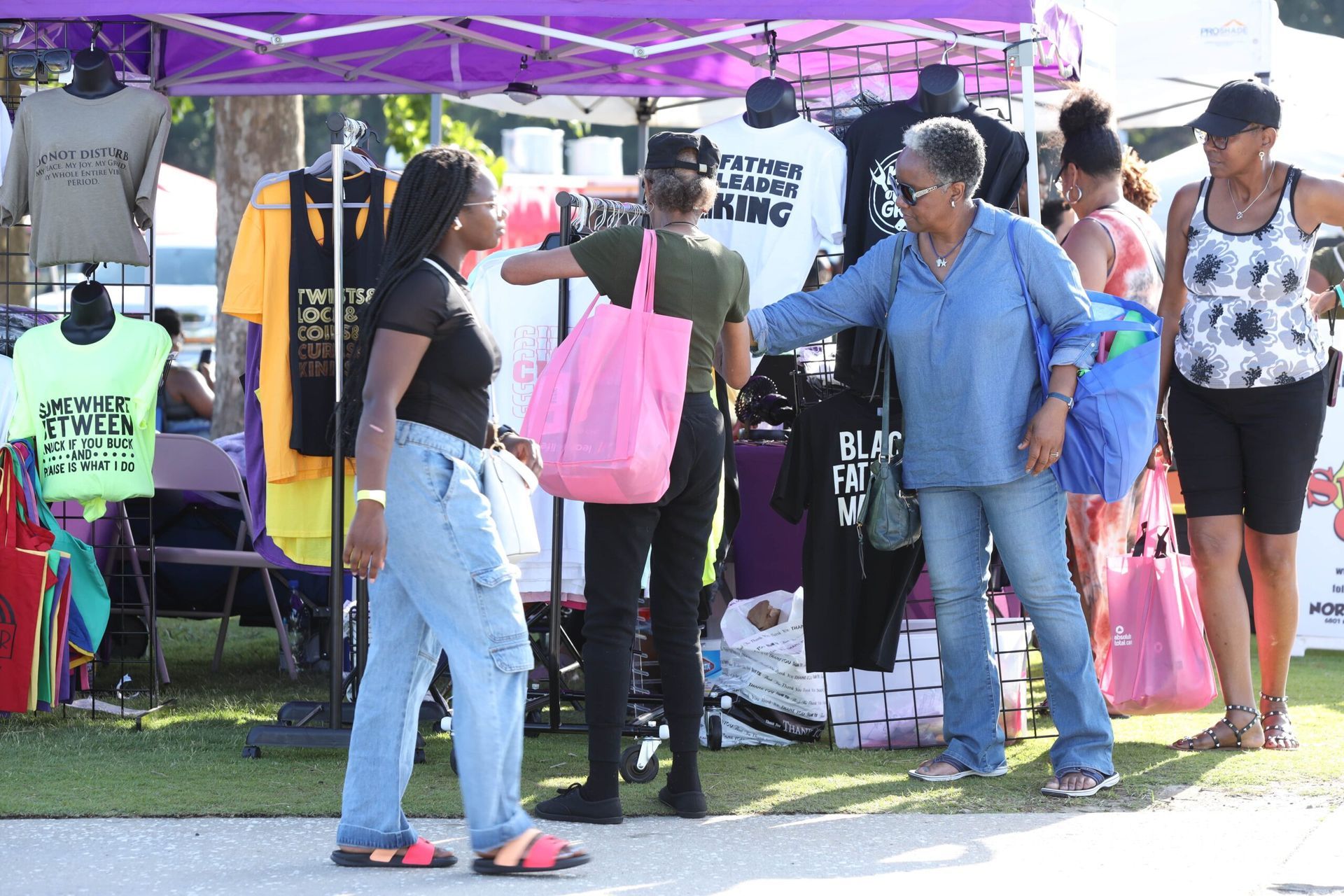 People gathered under a purple canopy at an outdoor event, with tents, tables, and bags on a grassy area.
