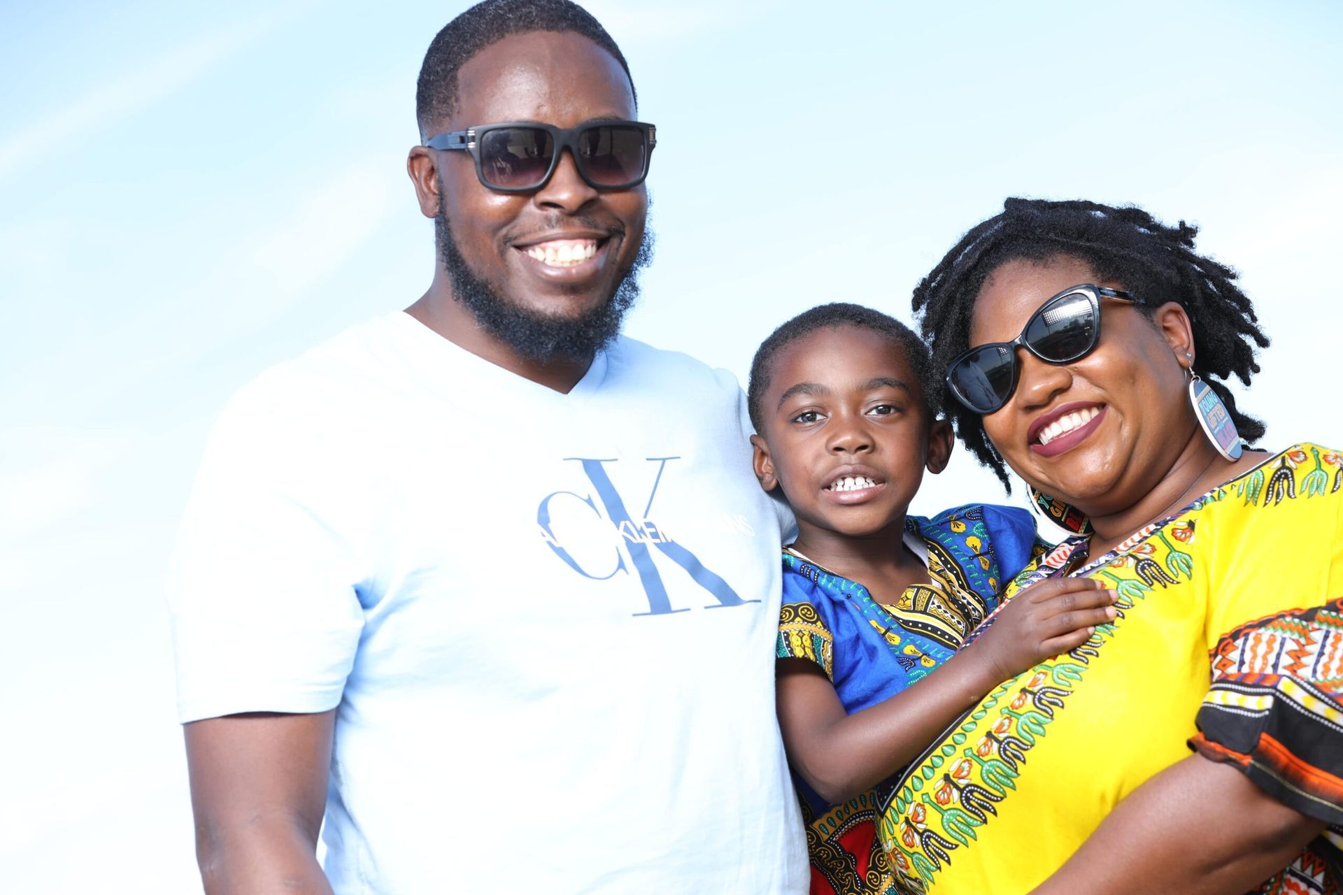 Three smiling people outdoors, wearing sunglasses and colorful clothing against a bright sky