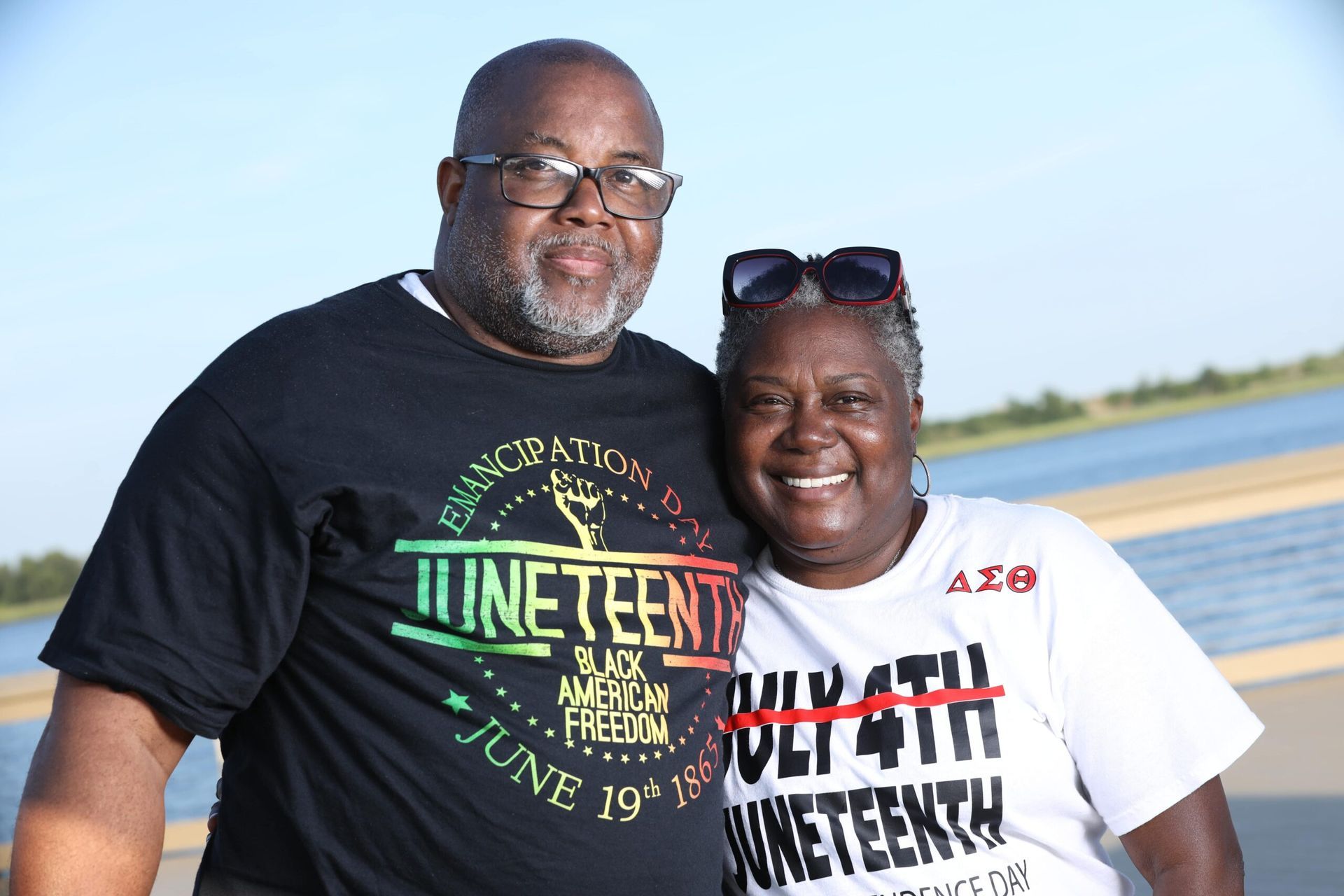 Smiling couple standing outdoors by the water, wearing Juneteenth shirts.