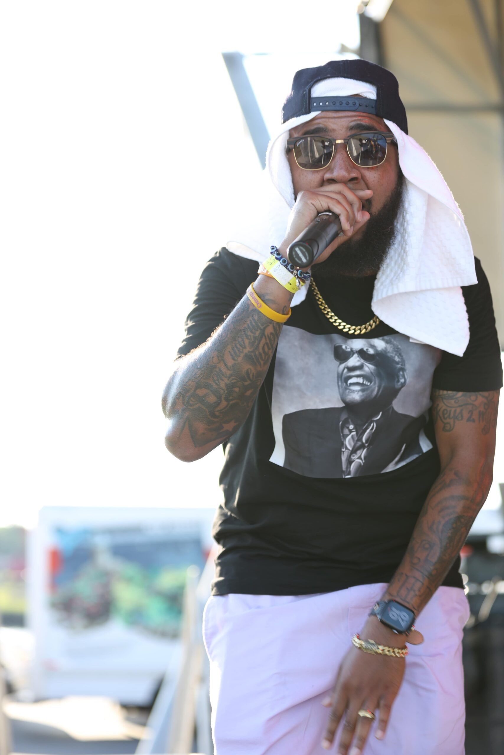 Person performing outdoors in sunglasses, black T-shirt, white pants, and towel over cap, holding a microphone.