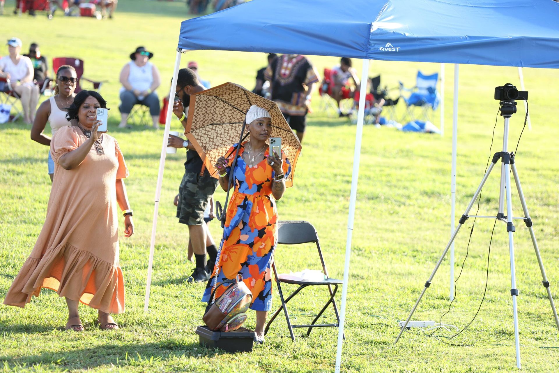 Two people under a blue canopy in a sunny field, one holding a patterned umbrella and one walking nearby