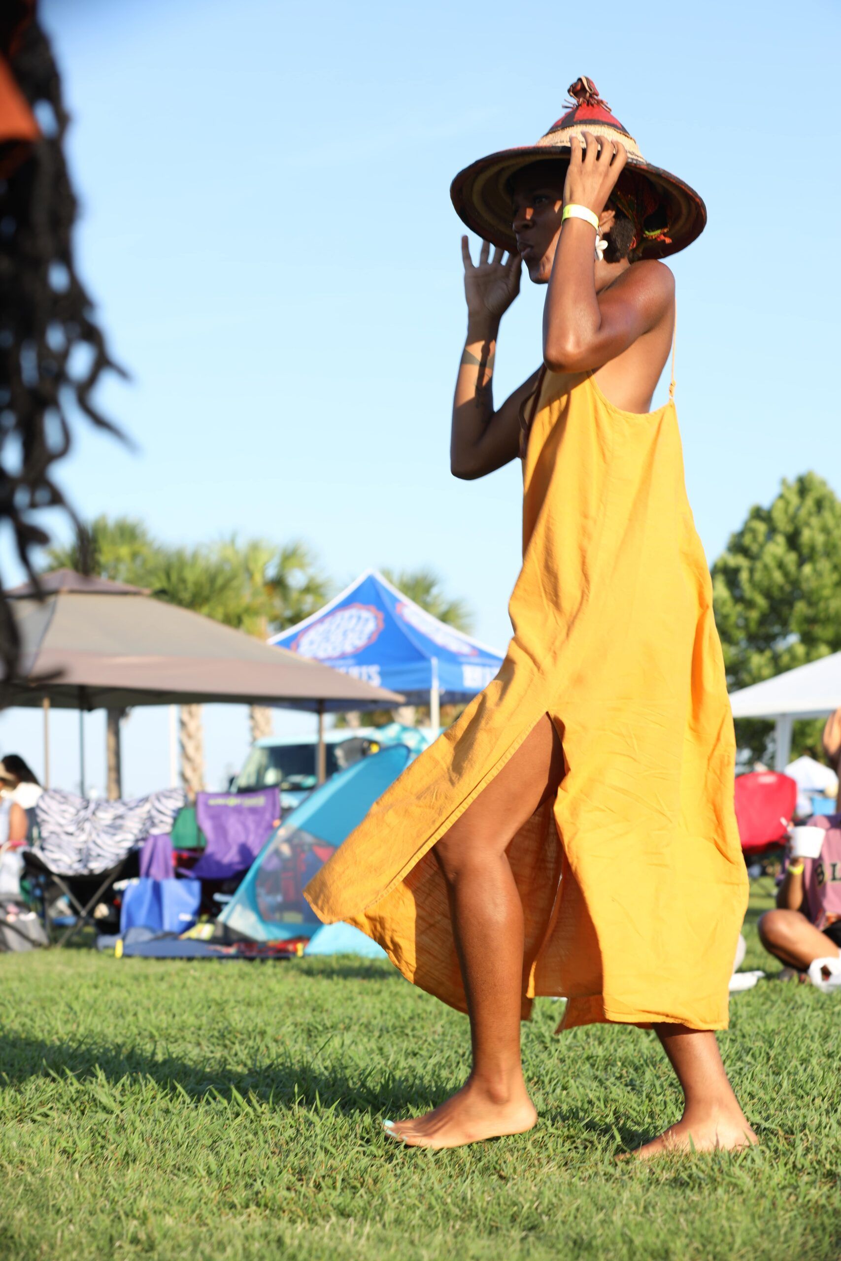 Person in a yellow dress and sun hat walking on grass at an outdoor event.