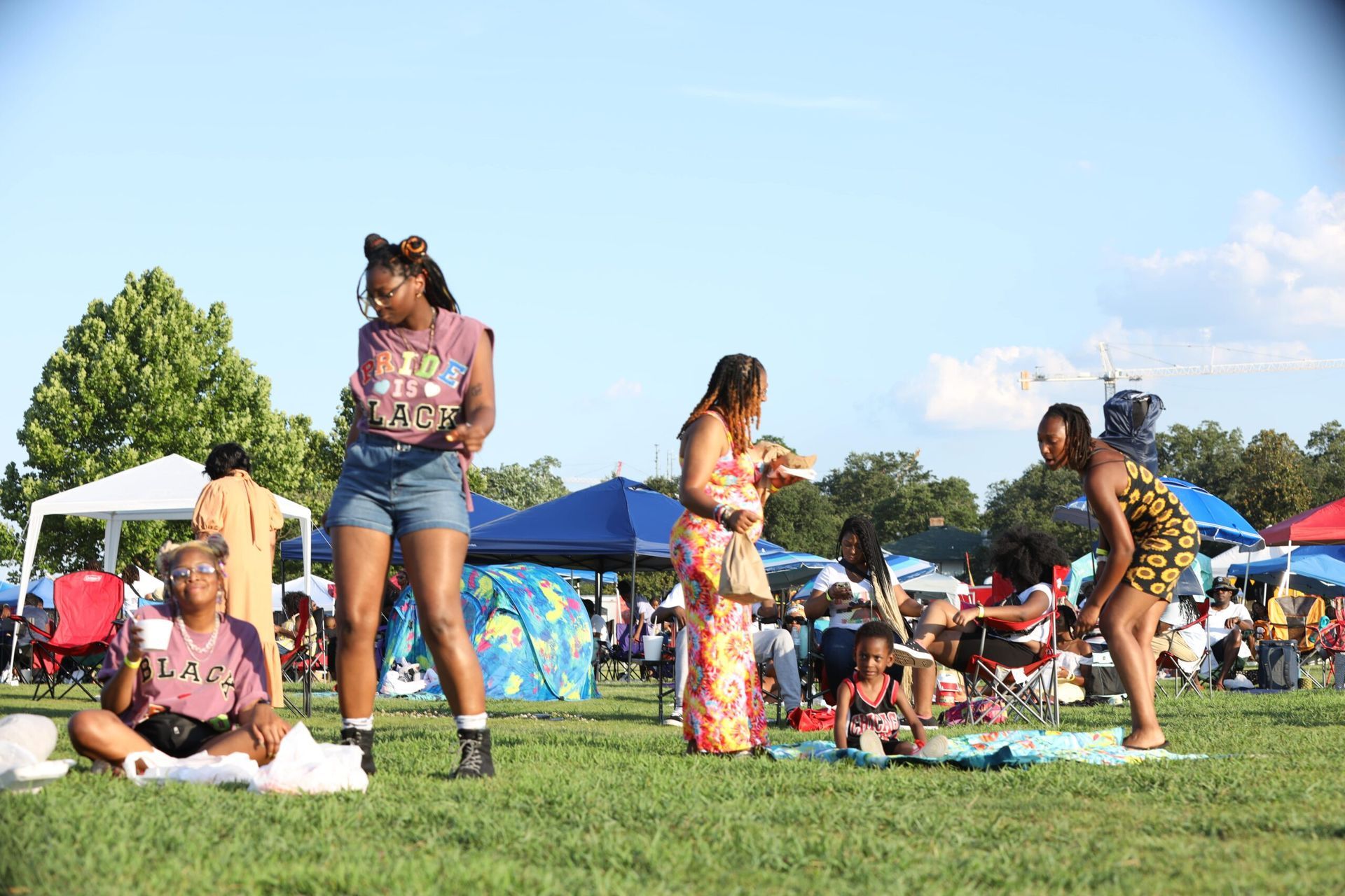 People standing and sitting on a grassy field at an outdoor festival under blue tents on a sunny day