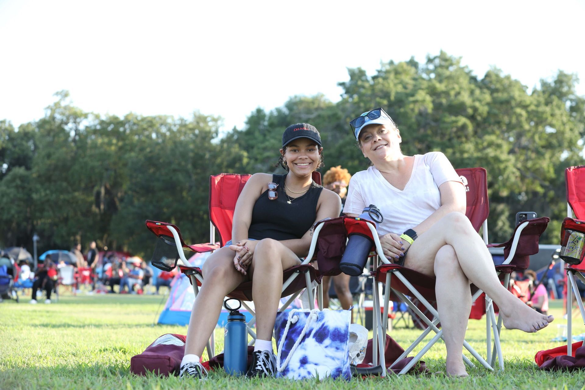 Two people smiling in folding chairs on a grassy field at an outdoor event