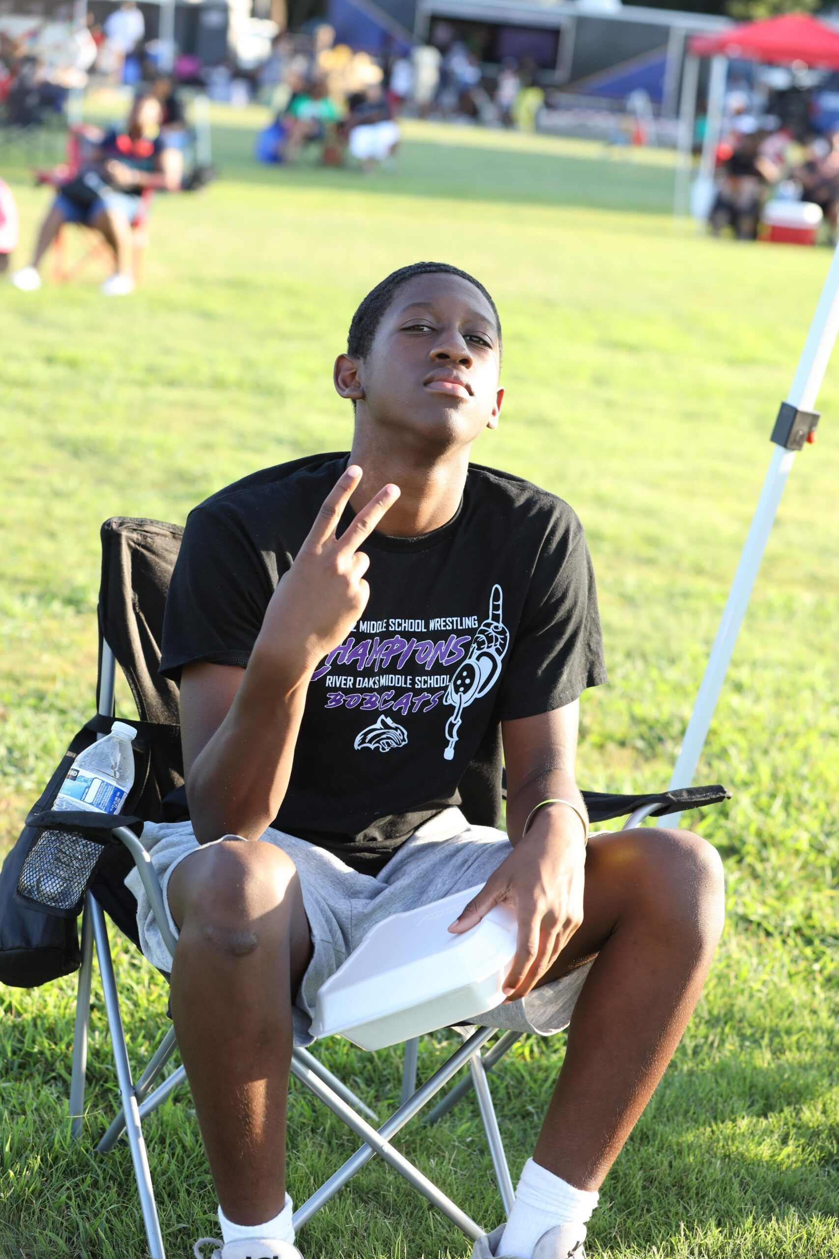 Person seated on a folding chair outdoors, wearing a black T-shirt and white shorts, holding up two fingers.