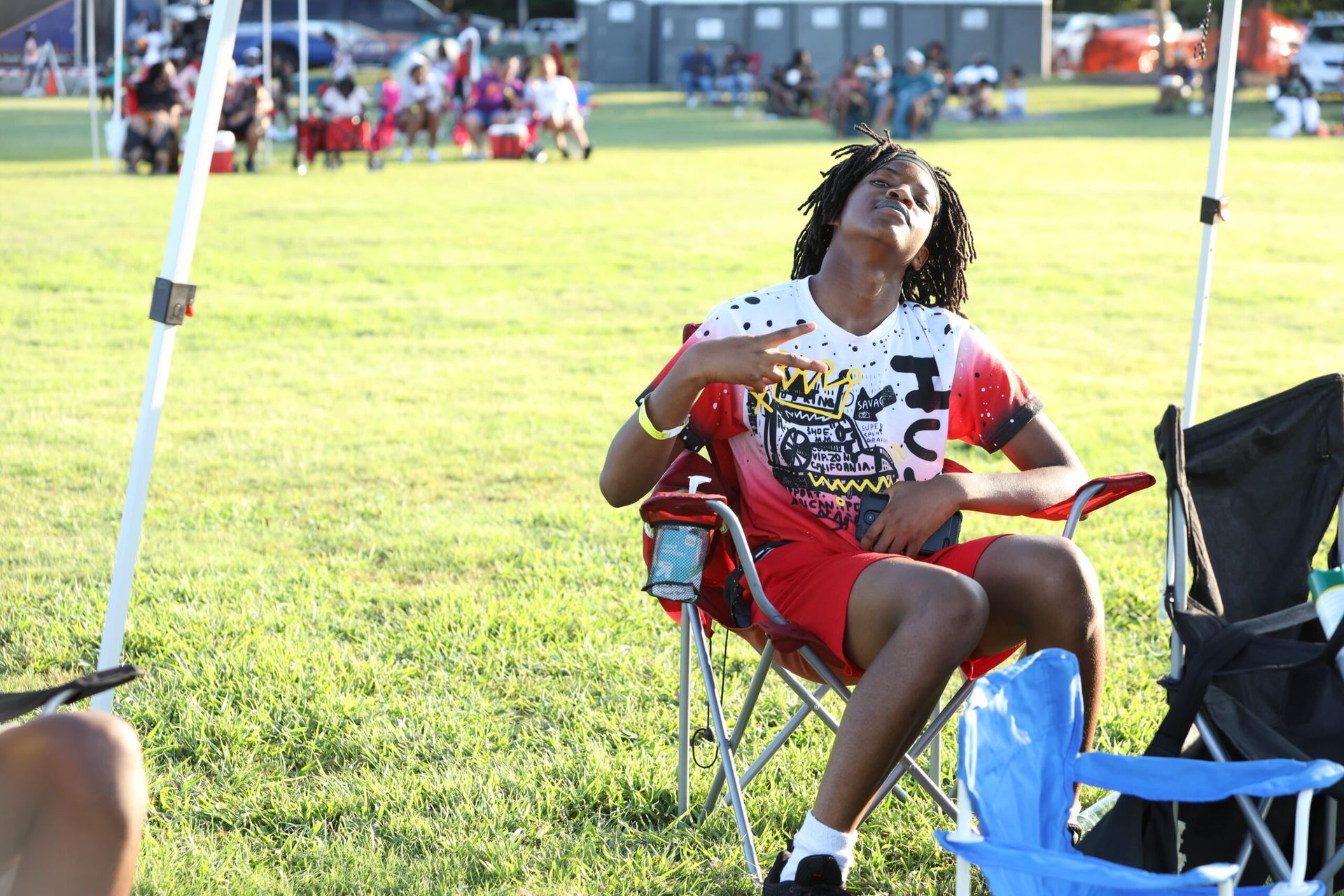 Person reclining in a red folding chair on a sunny grassy field, with tents and people in the background