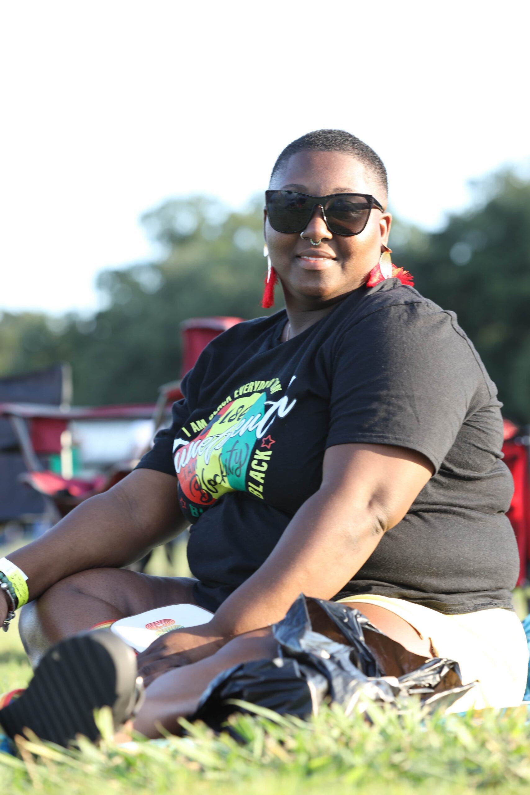 Woman sitting on grass outdoors, wearing sunglasses and a black graphic T-shirt.