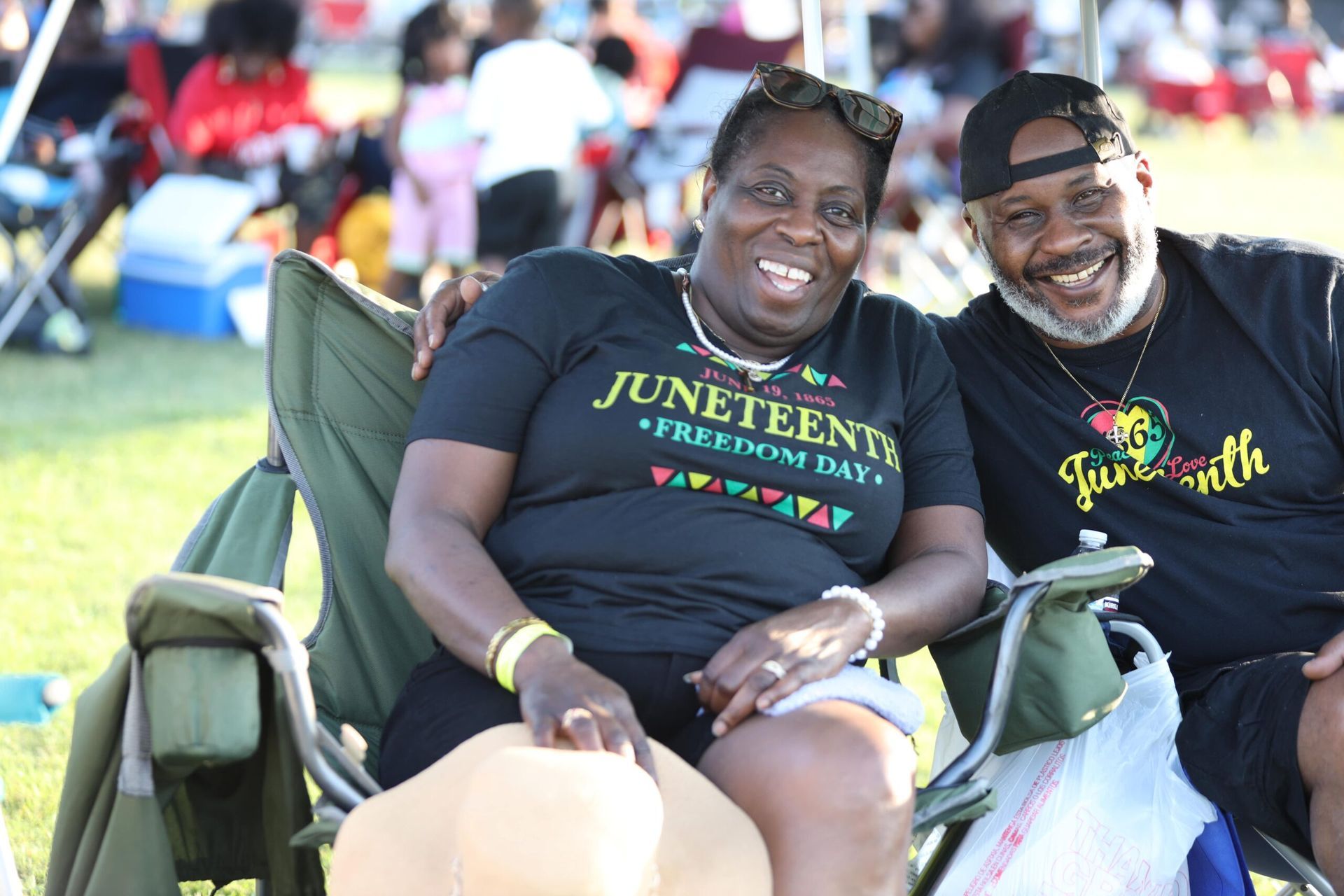 Two people smiling in lawn chairs at an outdoor Juneteenth celebration, wearing matching black shirts.