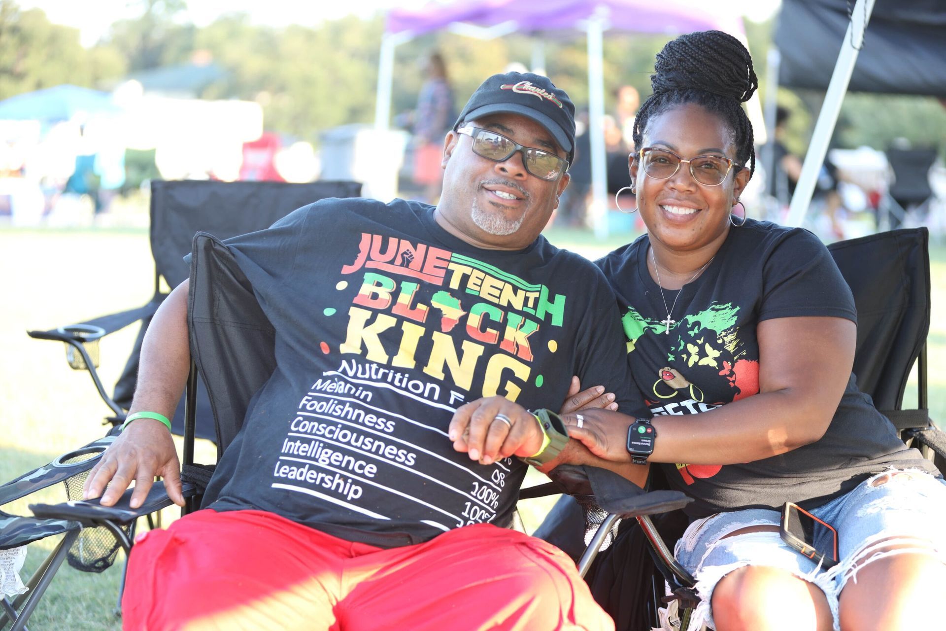 Two smiling people seated outdoors, wearing black T-shirts and sunglasses on a bright sunny day.