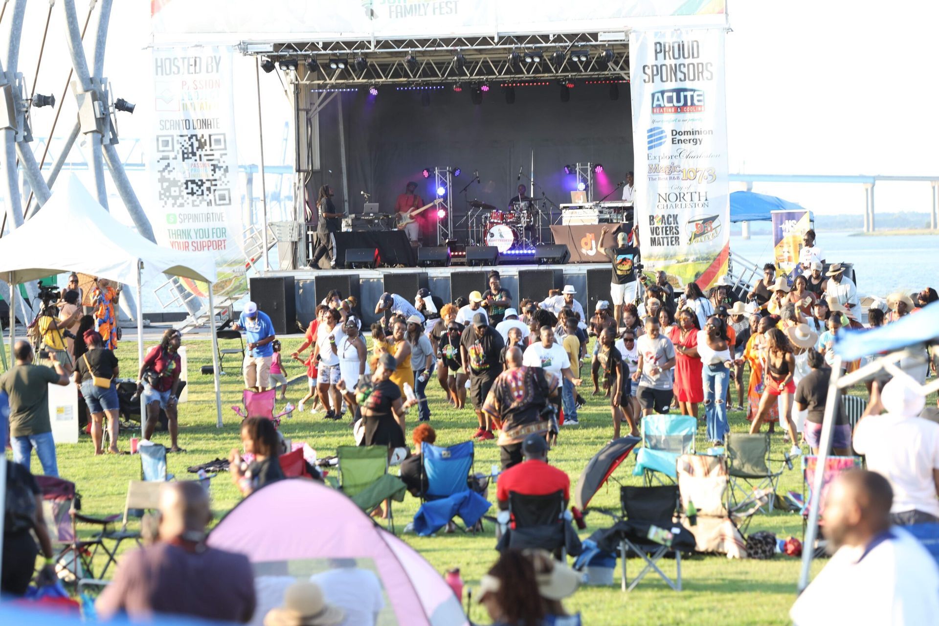 Outdoor music festival with a crowd on the grass in front of a stage, tents and people in colorful clothes.