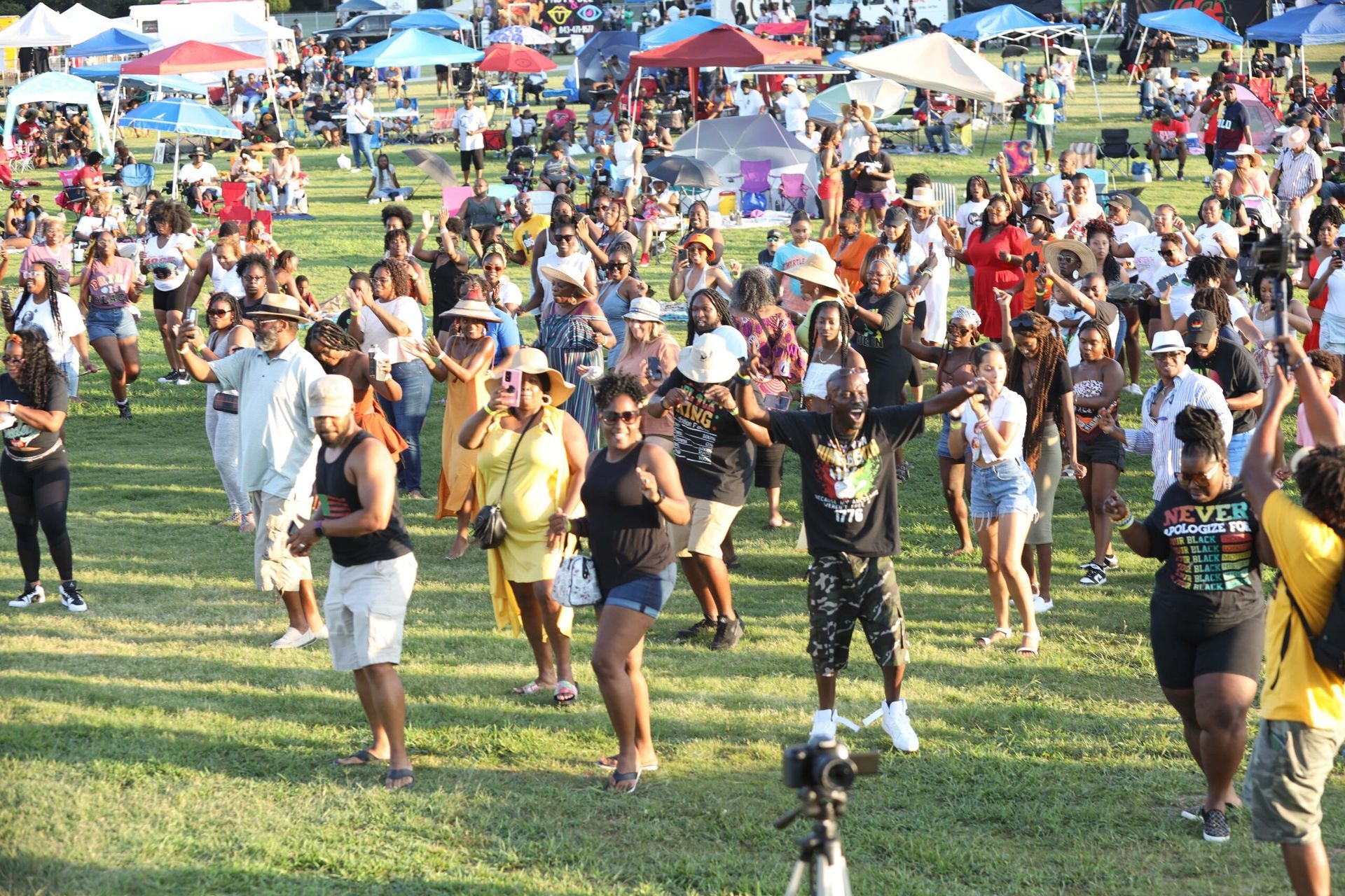 Crowd dancing and walking on a grassy field at an outdoor festival with colorful tents in the background