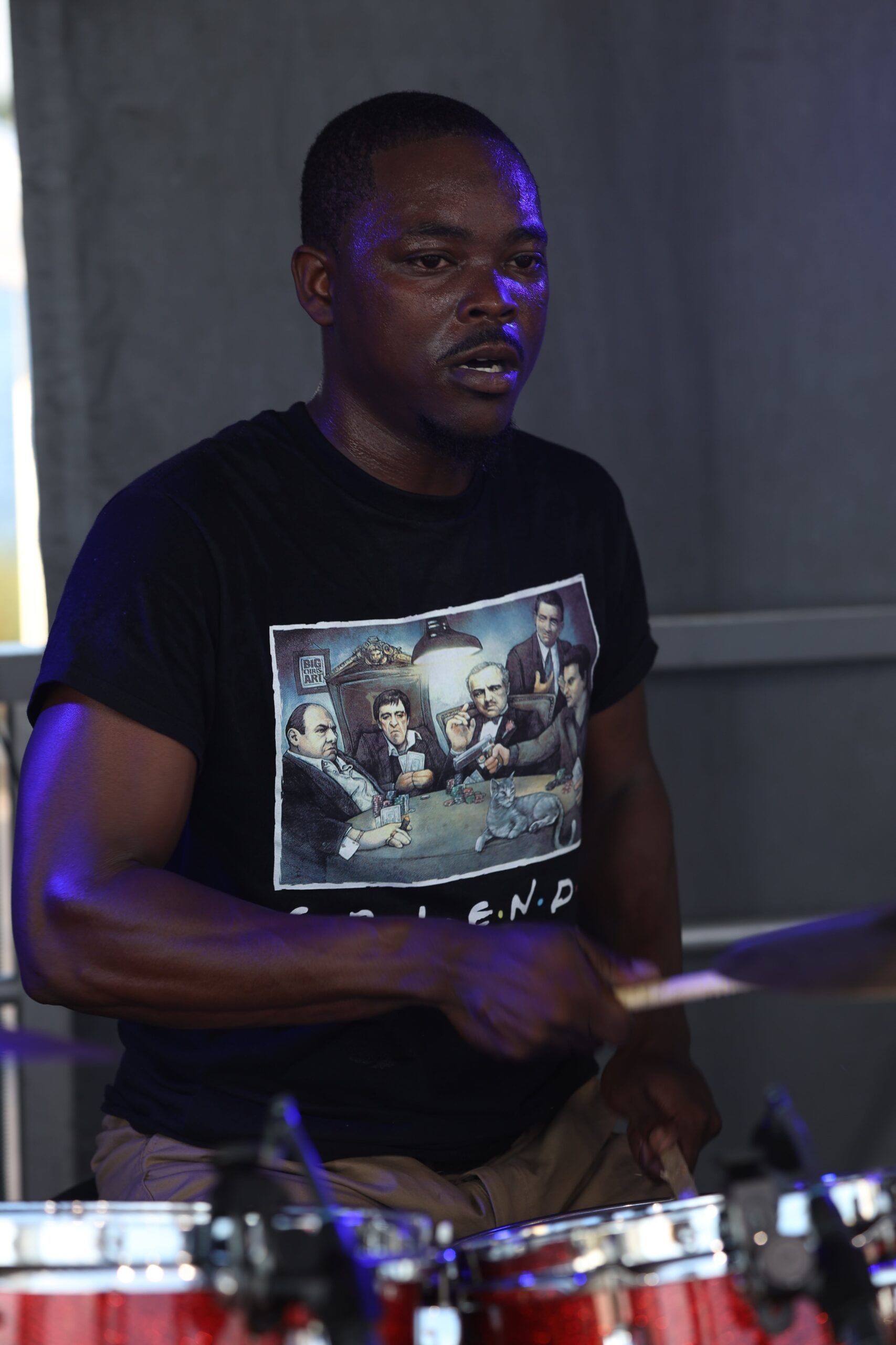 Man playing drums on stage in a black T-shirt, lit by purple stage lights.