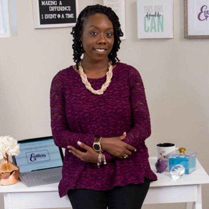 Woman in a purple top standing in a bright office, leaning against a white desk with framed wall art behind her.