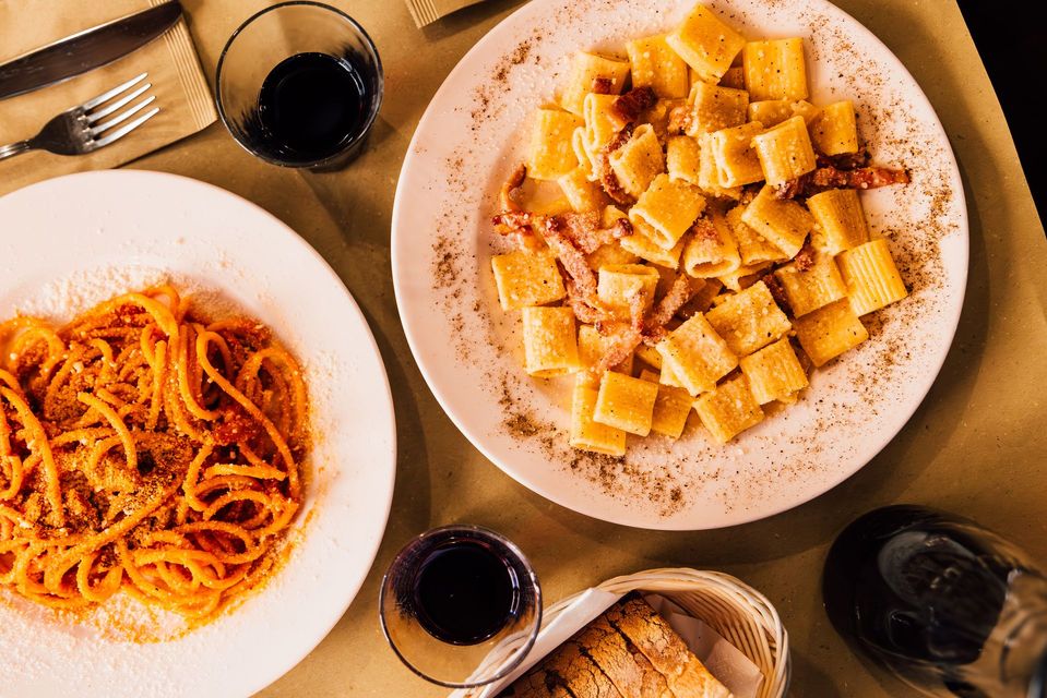 Due piatti di pasta, uno di spaghetti al sugo rosso, uno di rigatoni con pancetta, bicchieri di vino e pane su un tavolo.