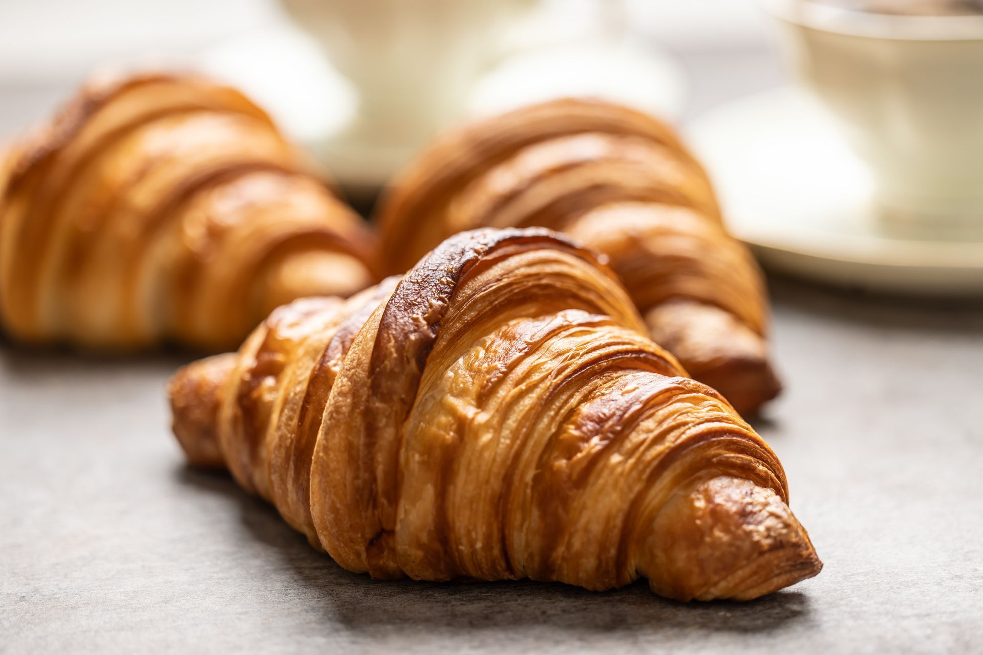 Tre croissant dorati su una superficie grigia, con tazze da tè sfocate sullo sfondo.