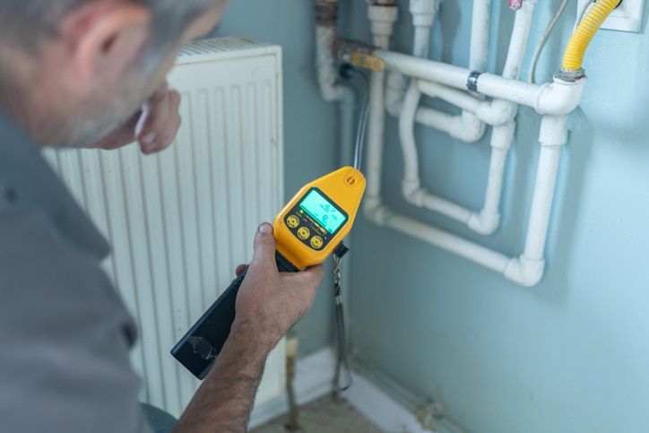 Technician using a yellow thermal camera to inspect white heating pipes near a radiator