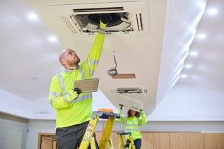 Technicians inspecting a ceiling air vent, one on a ladder reaching into an open duct