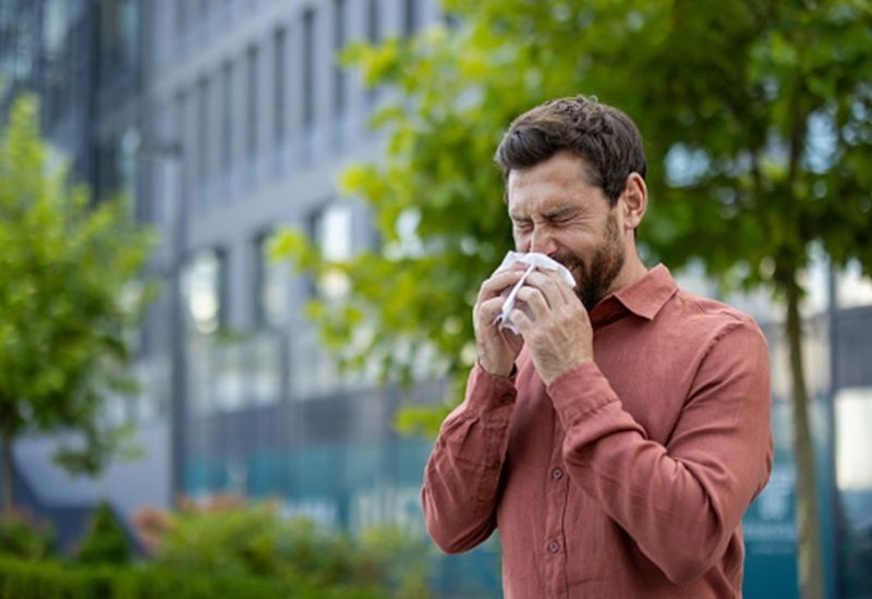 Man in a red shirt outdoors, blowing his nose with a tissue near a building and trees