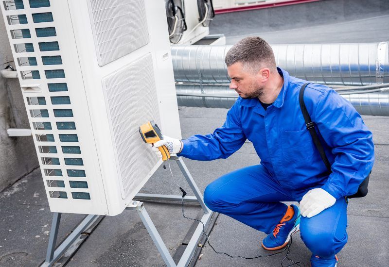 Technician in blue coveralls inspecting an outdoor HVAC unit with a handheld meter.