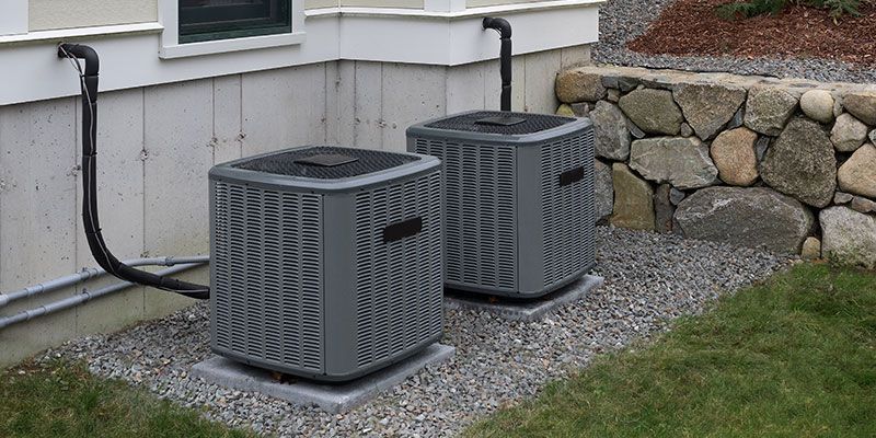 Two gray outdoor HVAC units beside a house, on gravel near a stone retaining wall and grass.