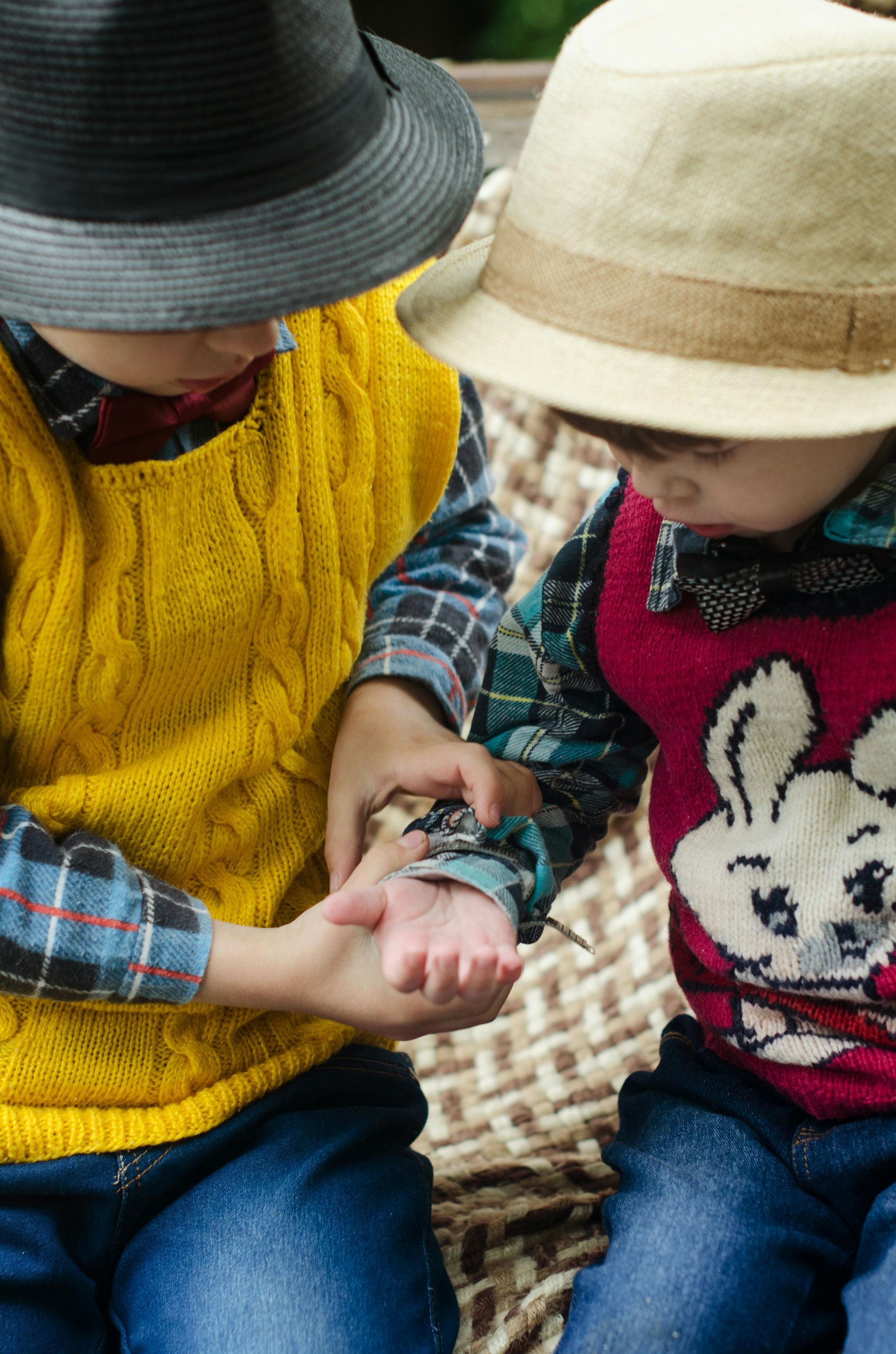Two young boys wearing hats and sweaters are sitting on a wicker chair.