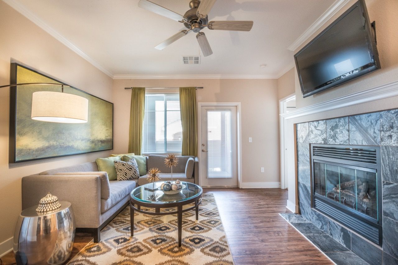 Cozy apartment living room with gray sofa, fireplace, wall-mounted TV, ceiling fan, and glass coffee table.