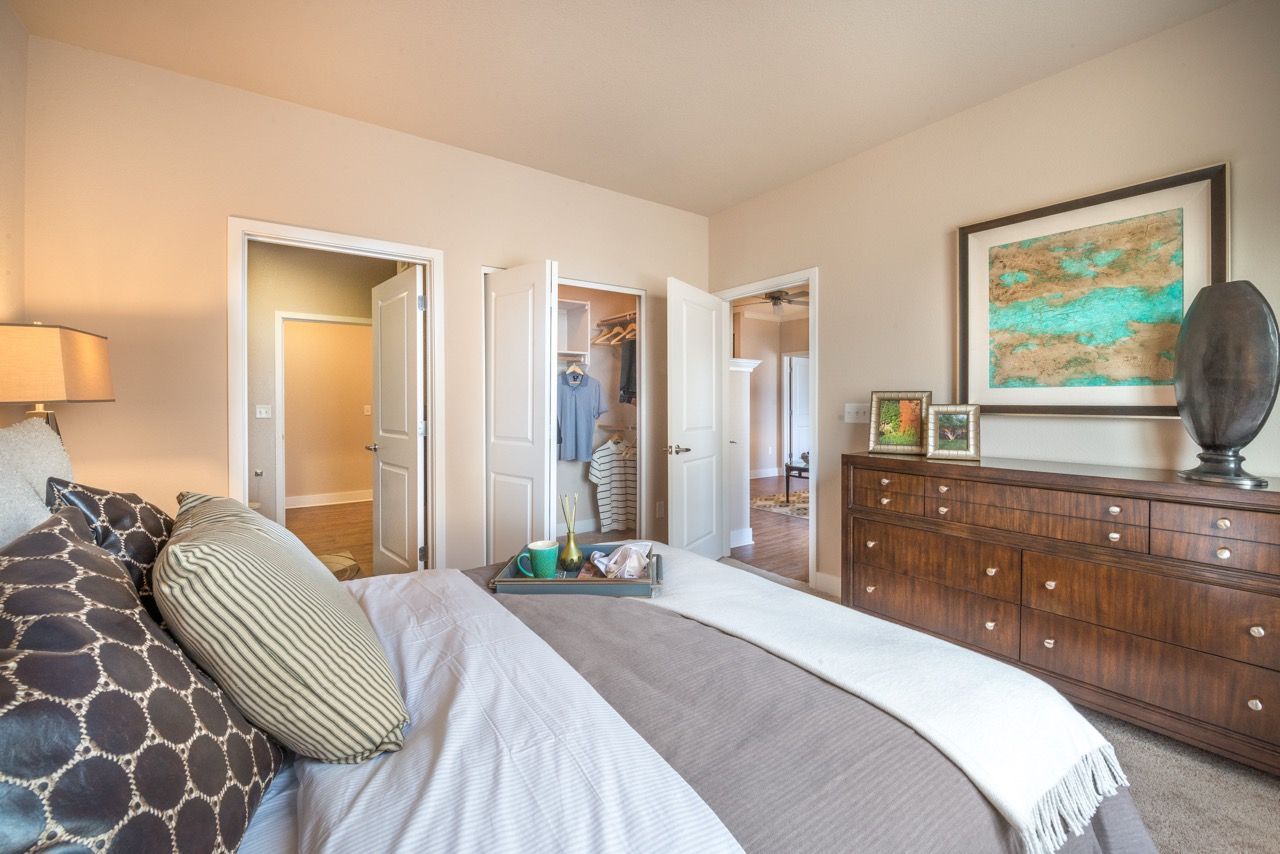 Furnished bedroom with a large bed, wood dresser, framed art, and open closet doors.