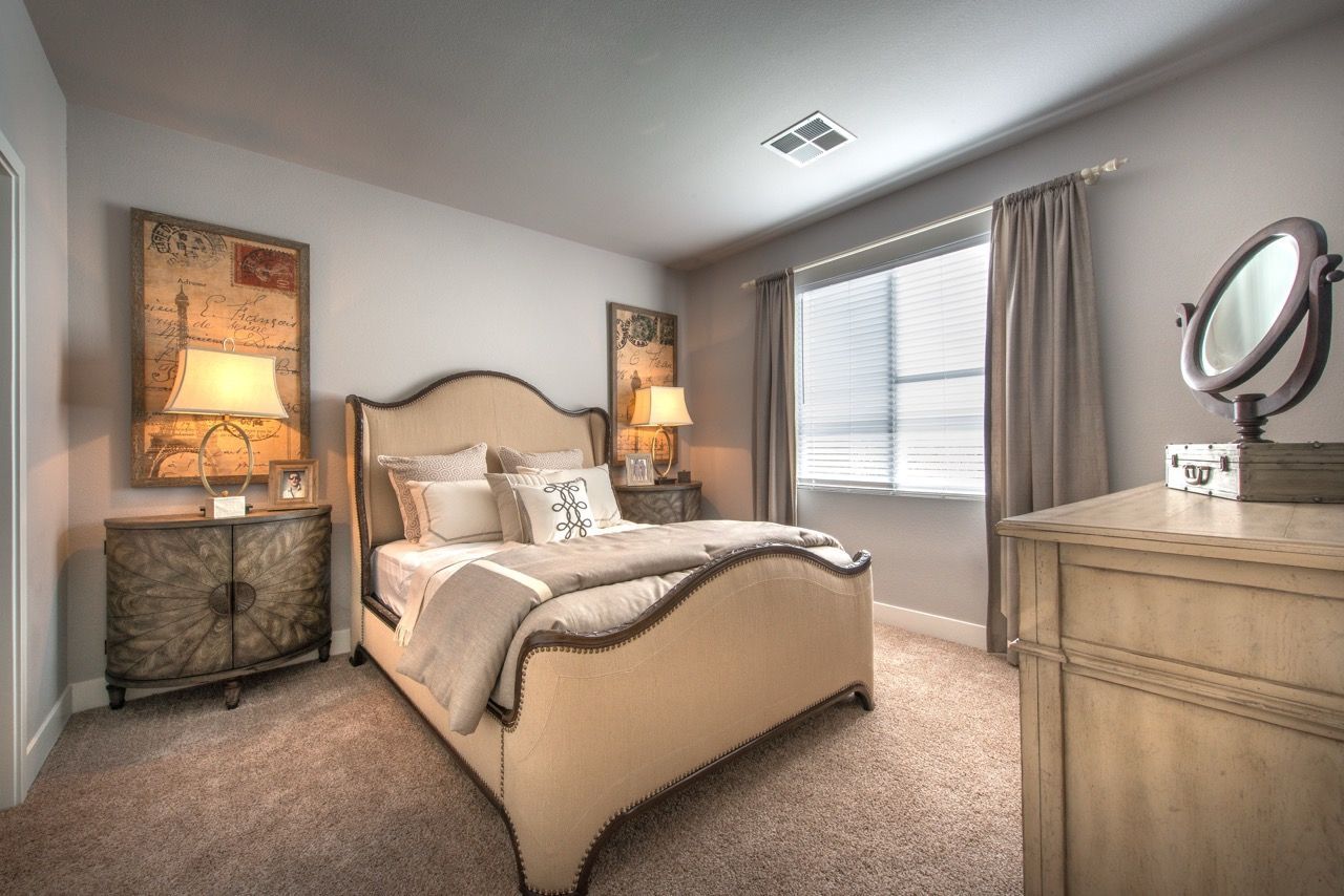 Bedroom in an apartment with an upholstered beige bed, two nightstands, a dresser, and a window with curtains.