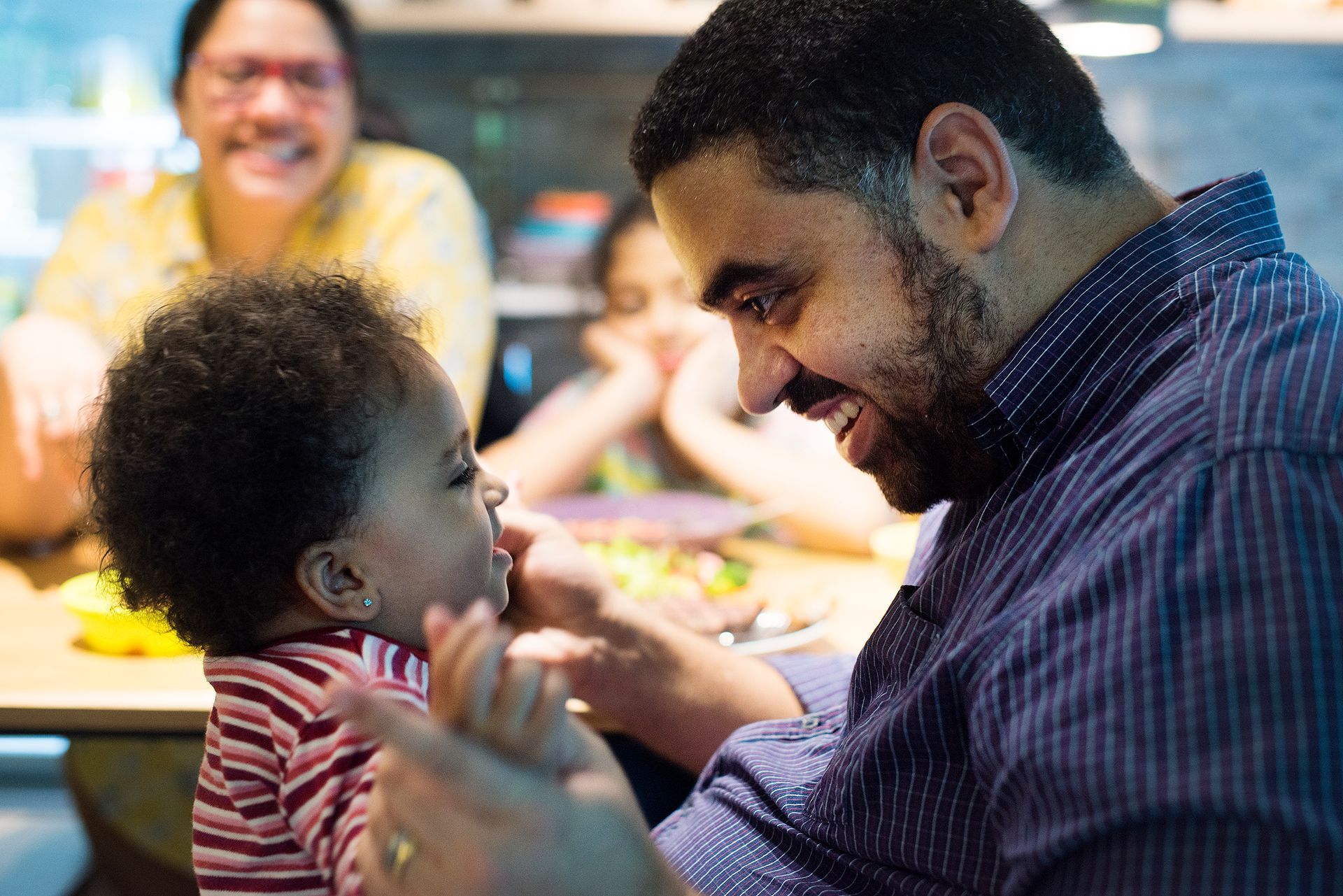 Man smiles at baby, reaching out. Others in background: woman, child. Indoors, bright.