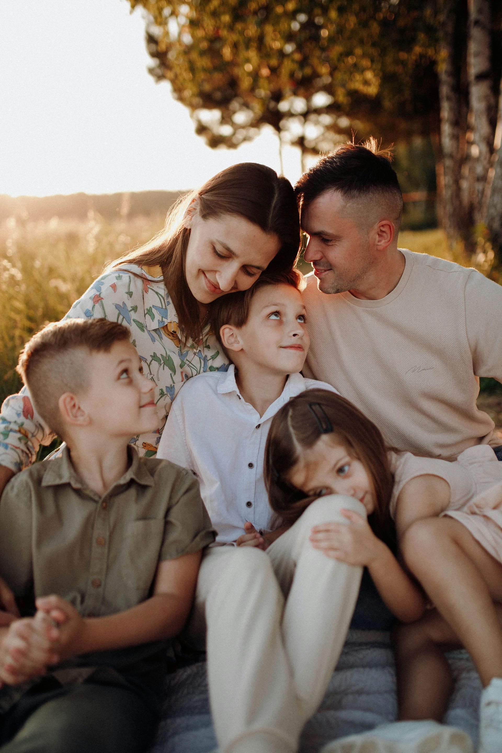 Family of five smiles while sitting outdoors in a field during golden hour.