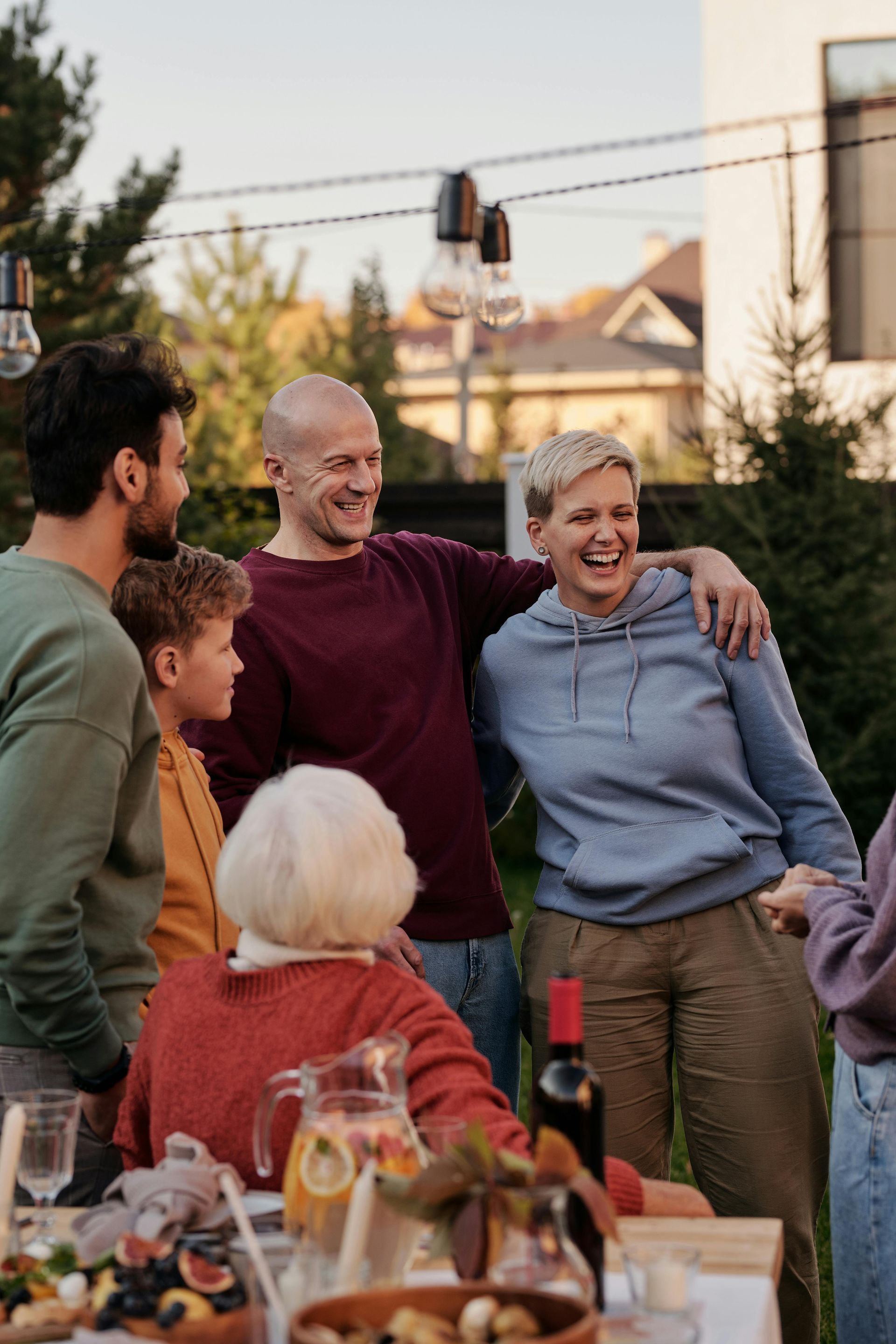 Group of people smiling, gathered around a table outdoors. Festive lighting above.
