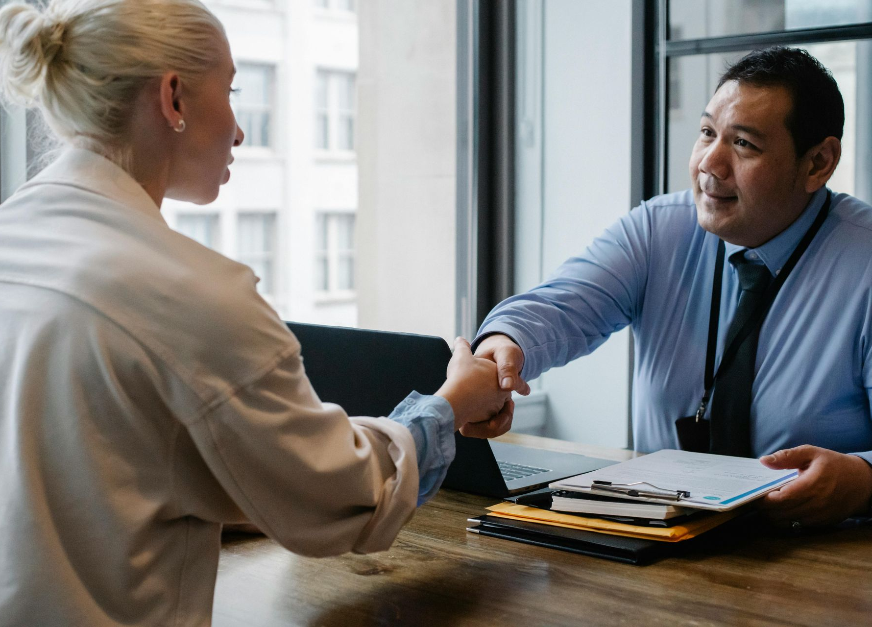 Woman and man shaking hands at a desk, likely in an office, with documents and a window in the background.